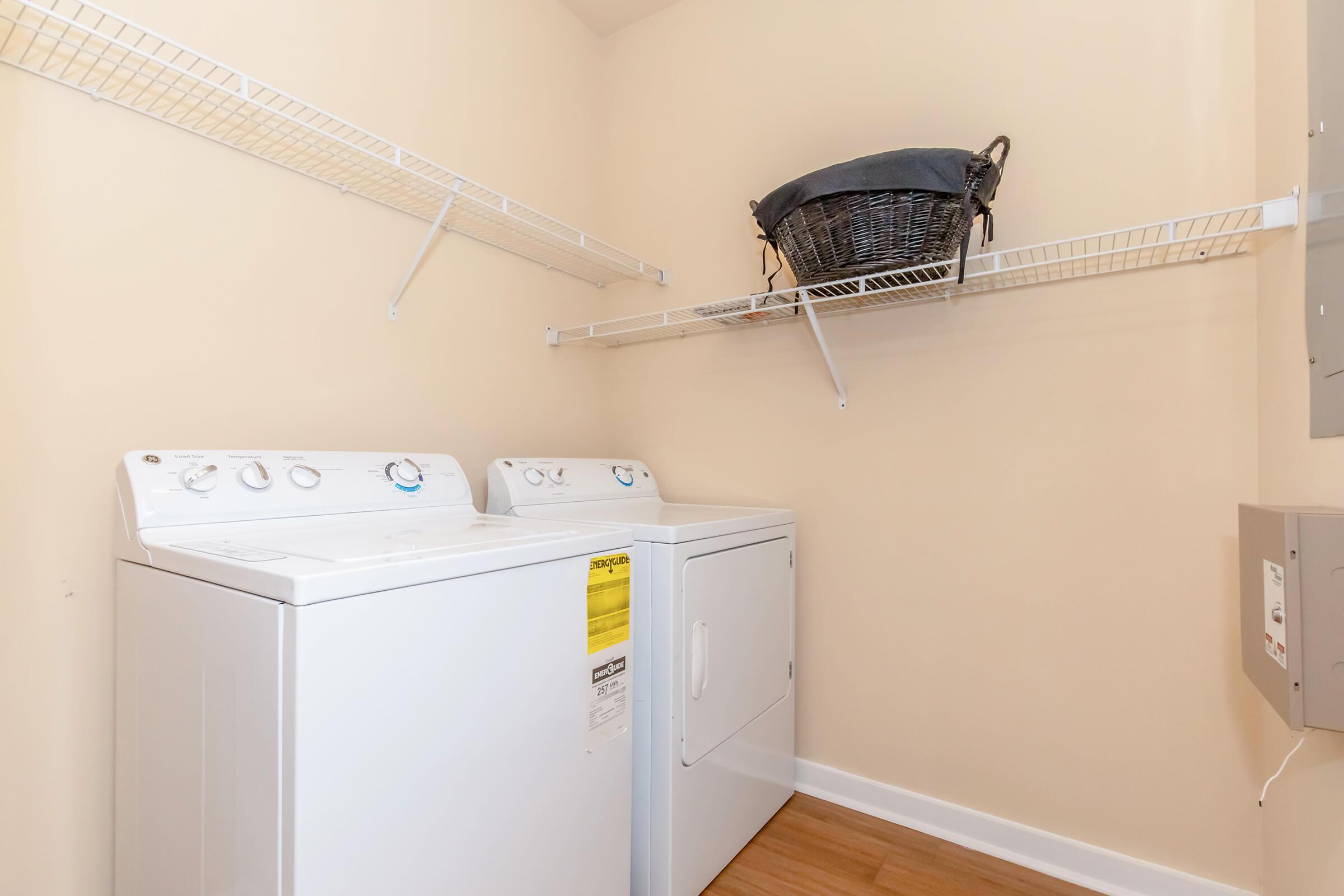A clean laundry room featuring a white washer and dryer side by side. Shelving above holds a woven laundry basket. The walls are a light beige, and the floor is a light hardwood. The space is well-organized and tidy, suitable for washing and drying clothes.