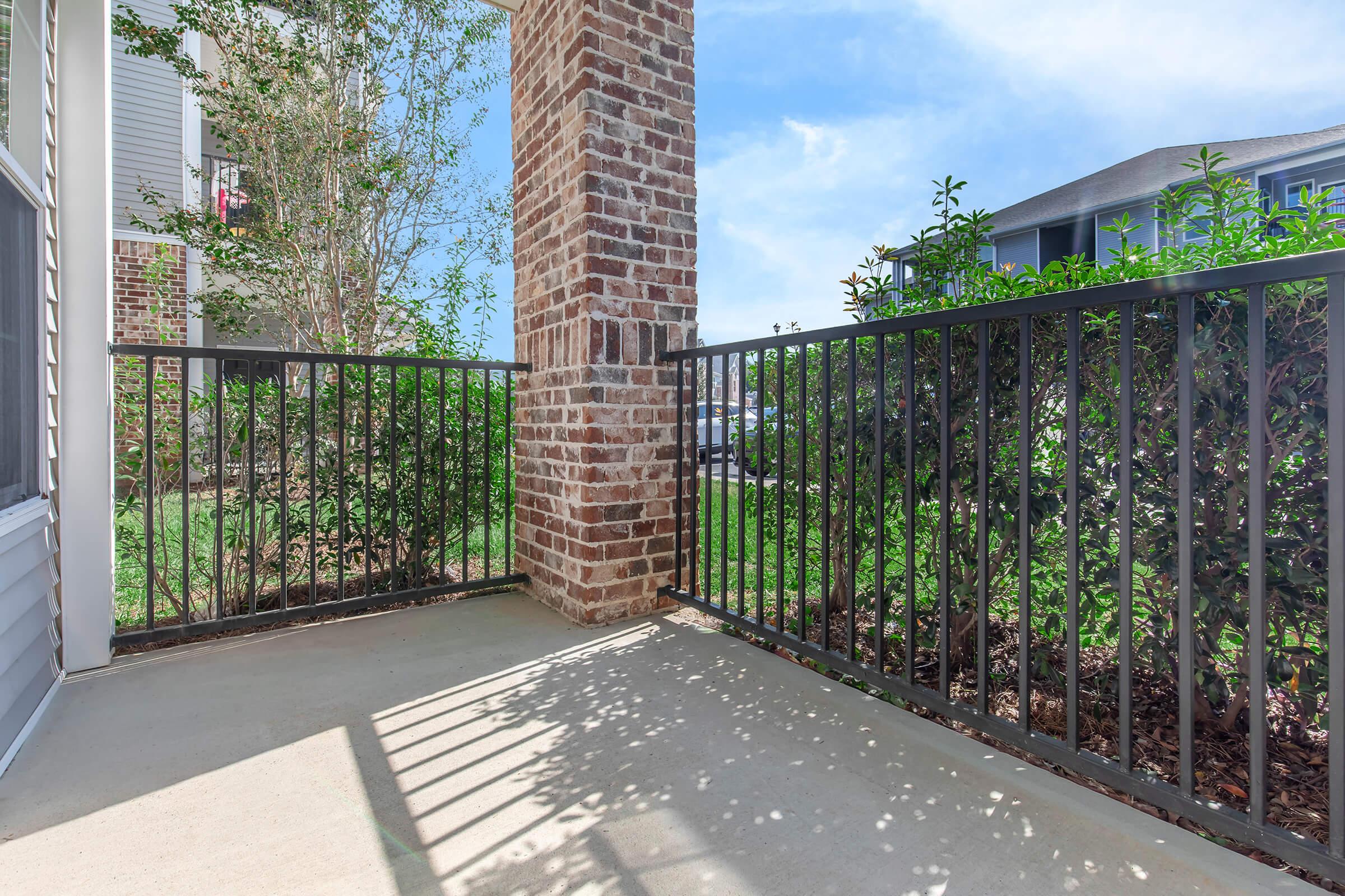 A corner balcony with a brick column, featuring a black metal railing. Lush green bushes are visible on one side, while the other side opens up to a grassy area. The space is well-lit with sunlight casting shadows on the concrete floor. Clear blue sky in the background enhances the outdoor feel.