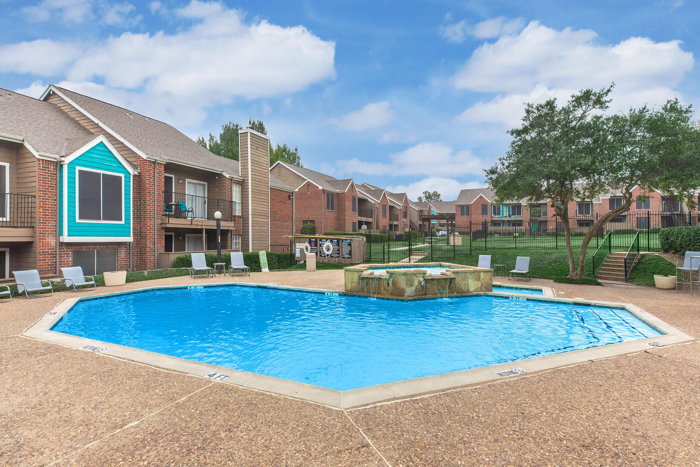 A well-maintained pool area with a sparkling blue pool surrounded by lounge chairs and a small stone fountain. In the background, there are two-story apartment buildings with a grassy area and trees, set against a partly cloudy sky. The space looks inviting and suitable for relaxation.