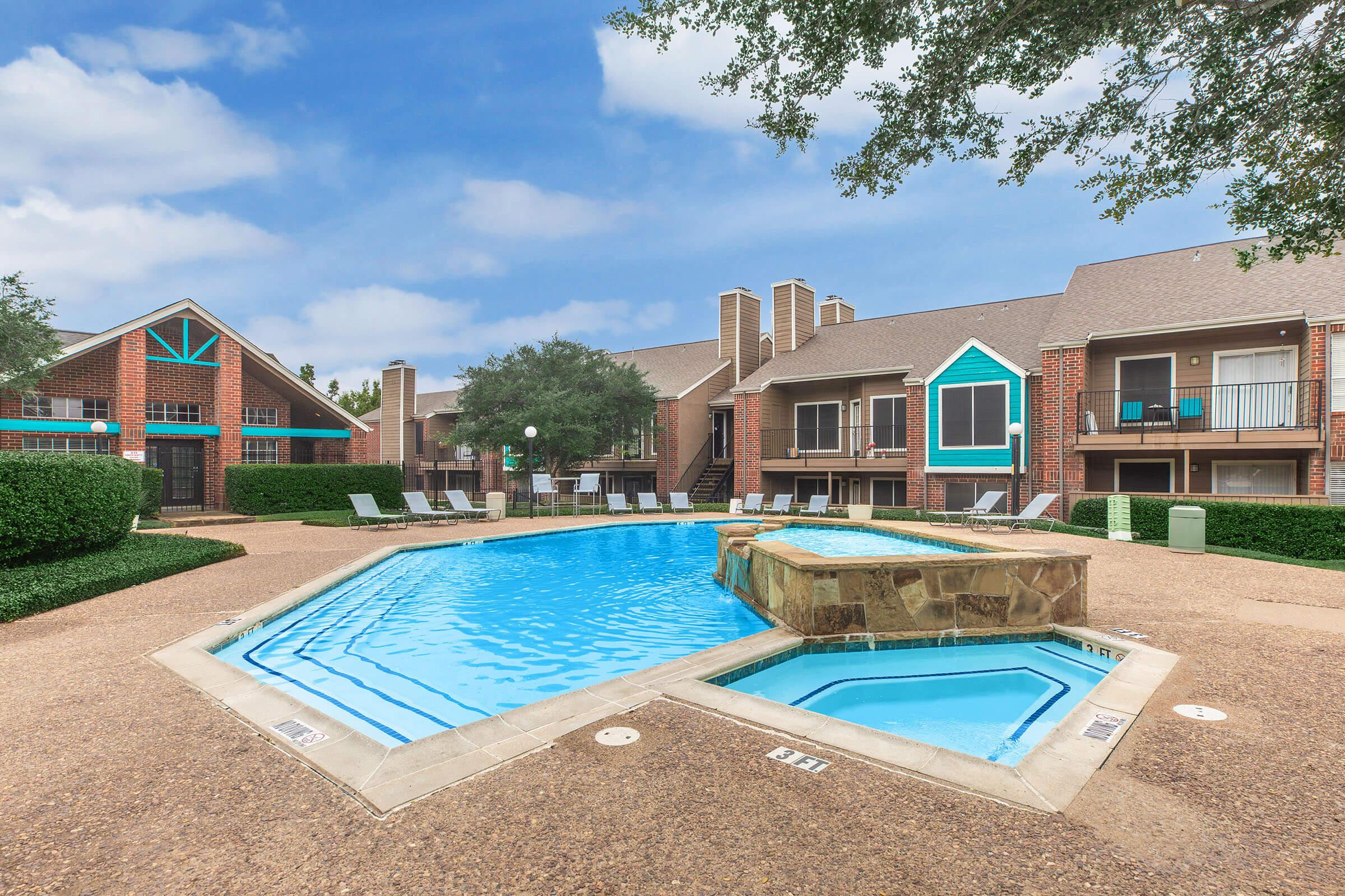 A clear swimming pool surrounded by lounge chairs, with a landscaped area featuring bushes and trees. In the background, there are buildings with balconies and a colorful facade, under a partly cloudy sky. The pool area provides a serene and inviting atmosphere for relaxation and leisure.