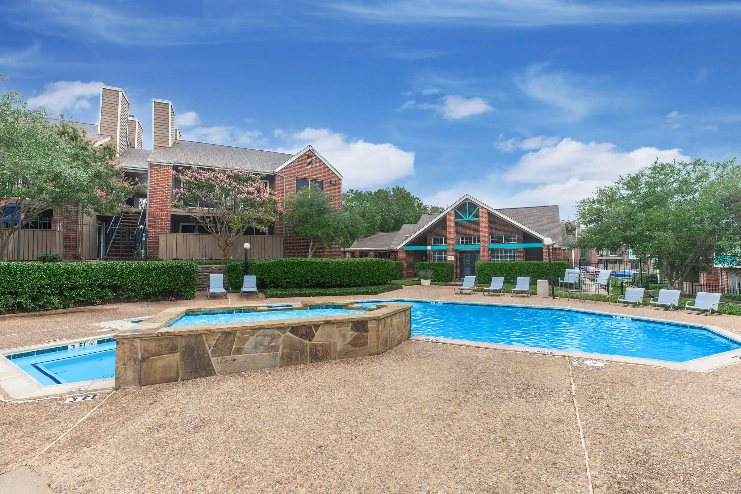 A serene outdoor swimming pool area surrounded by well-maintained landscaping, featuring lounge chairs and a nearby brick apartment complex under a clear blue sky. The pool has a shallow section and is framed by greenery, creating a relaxing and inviting atmosphere.