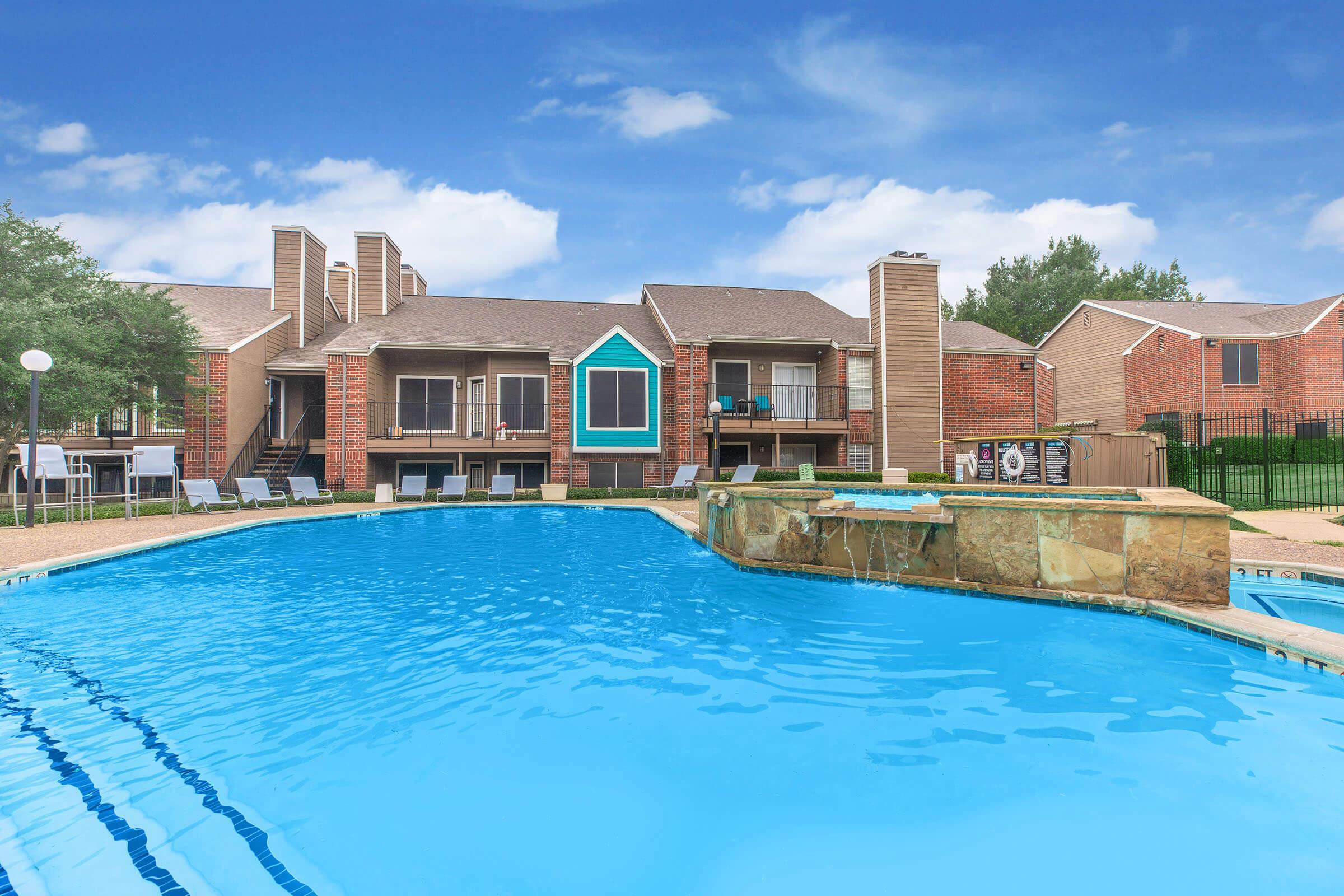 A clear blue swimming pool in the foreground with a stone edge, surrounded by lounge chairs. In the background, there are two-story apartment buildings with brick exteriors and balconies, set against a blue sky with a few clouds. Lush greenery adds a touch of nature to the scene.