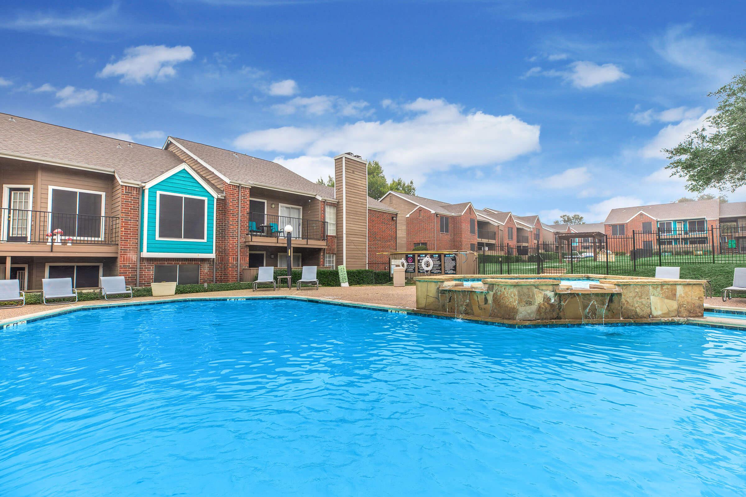 A serene swimming pool surrounded by lounge chairs, with brick apartment buildings in the background. The sky is clear with a few clouds, and a stone fountain is visible near the pool's edge. The scene conveys a relaxing outdoor atmosphere.
