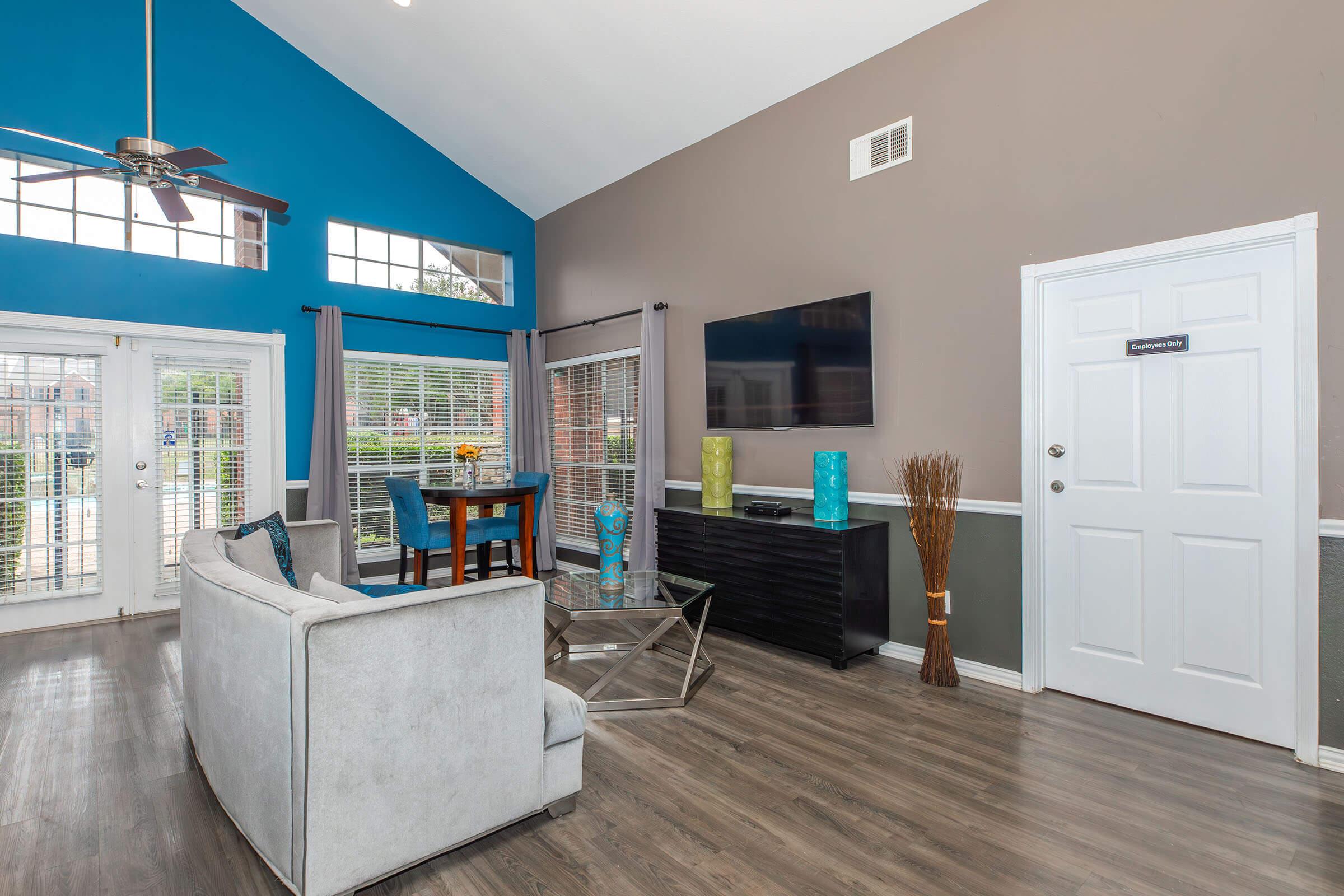Bright and modern living room featuring a light gray sofa, a glass coffee table, a dark wood TV stand, and decorative vases. Large windows allow natural light to fill the space, complemented by blue and tan walls. A dining table can be seen in the background, enhancing the open feel of the area.