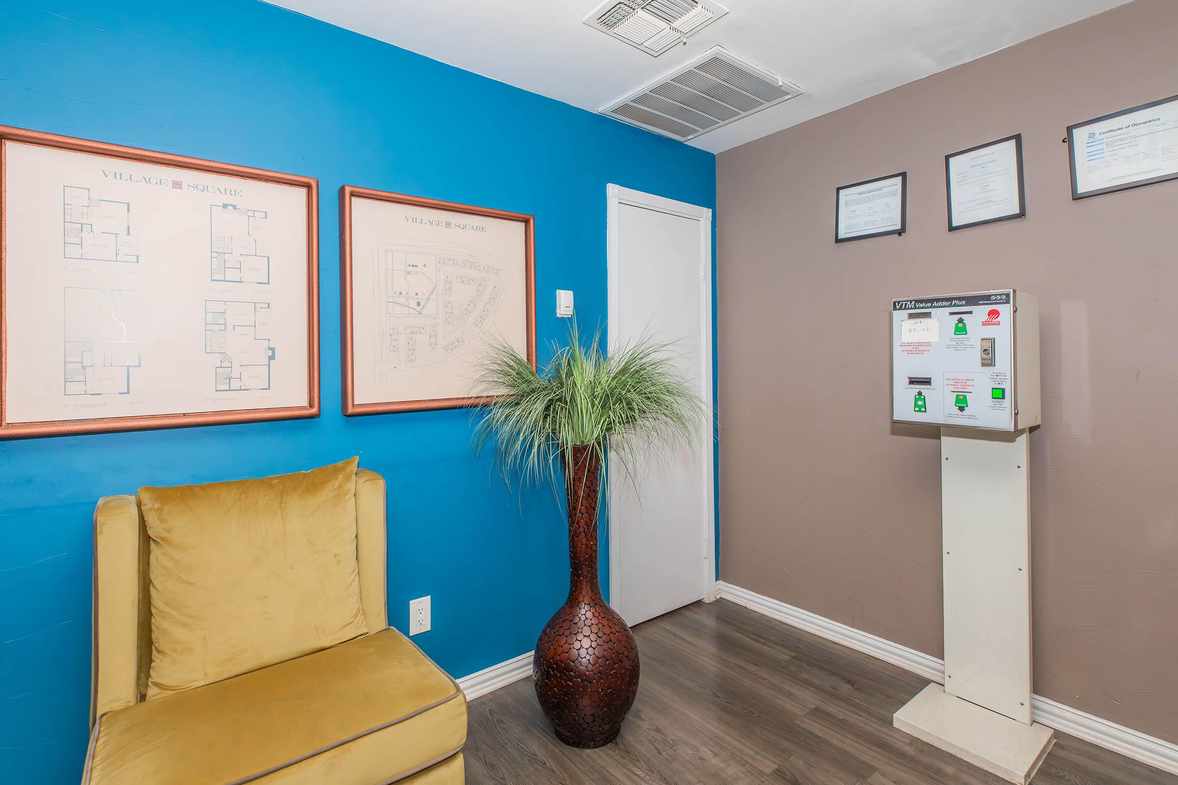 A cozy waiting area featuring a yellow armchair, blue and brown walls, framed floor plans, and a potted plant. A white information stand with various notices is present, along with several documents displayed on the wall above.