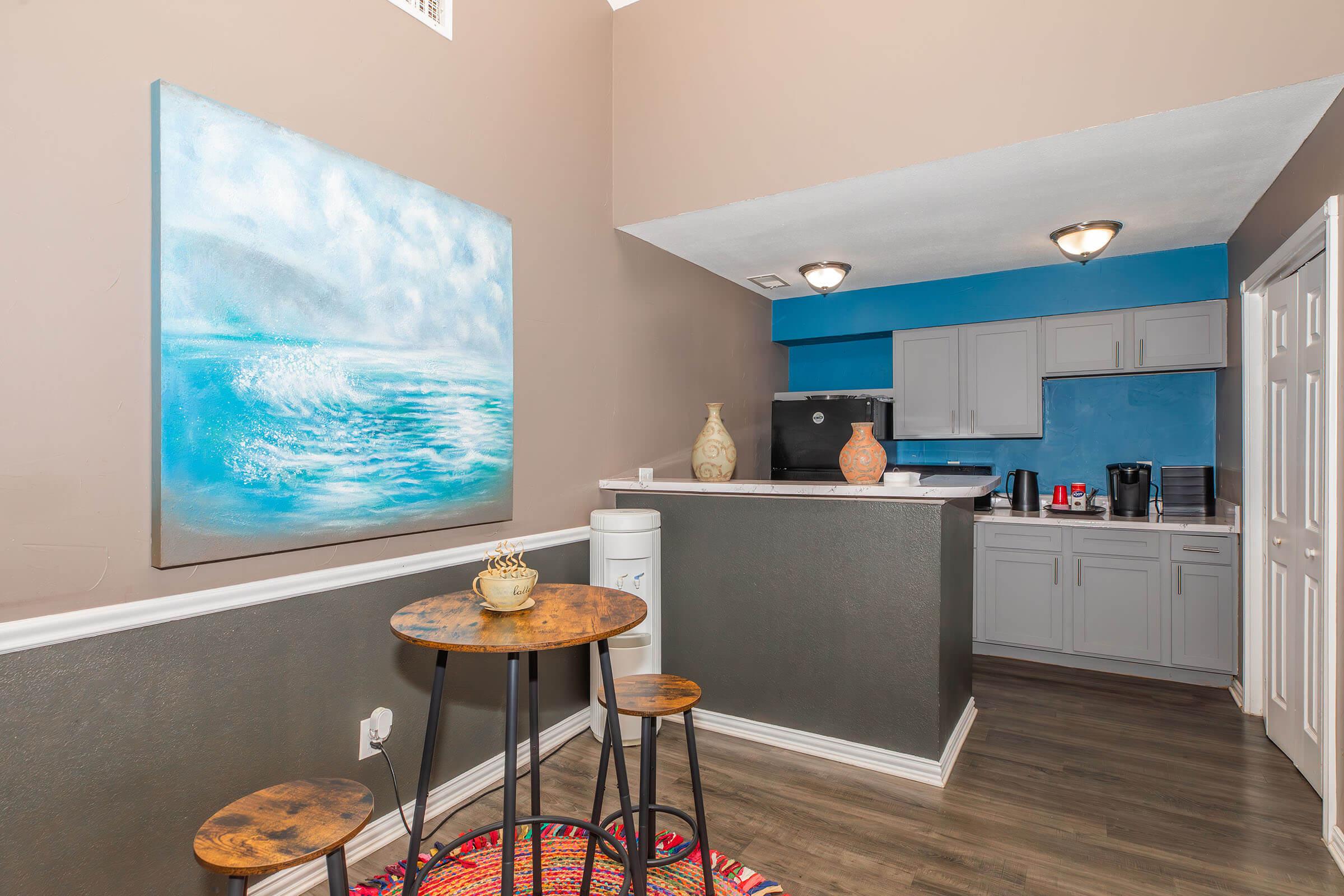 A modern kitchen and dining area featuring grey cabinets, a blue accent wall, and a circular wooden table with two stools. Artwork depicting a seascape hangs on the wall, and appliances are neatly arranged, with a colorful rug adding a touch of warmth to the space.