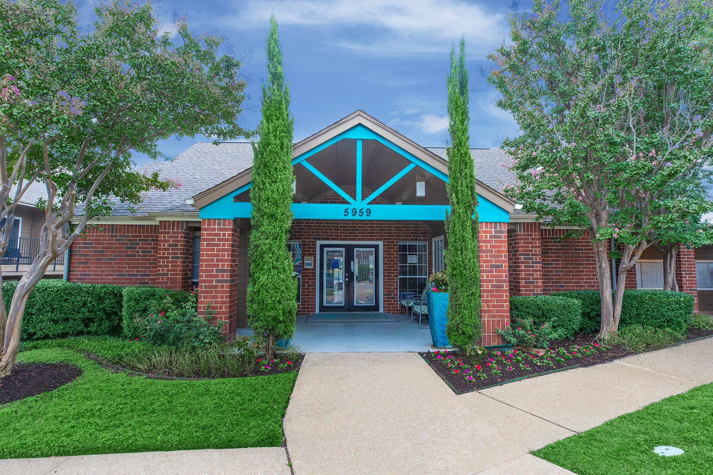 A modern building entrance featuring red brick walls and a blue-trimmed roof, surrounded by well-maintained greenery. The approach includes a concrete pathway lined with colorful flower beds and tall trees, creating a welcoming atmosphere. The building number "5959" is displayed prominently.