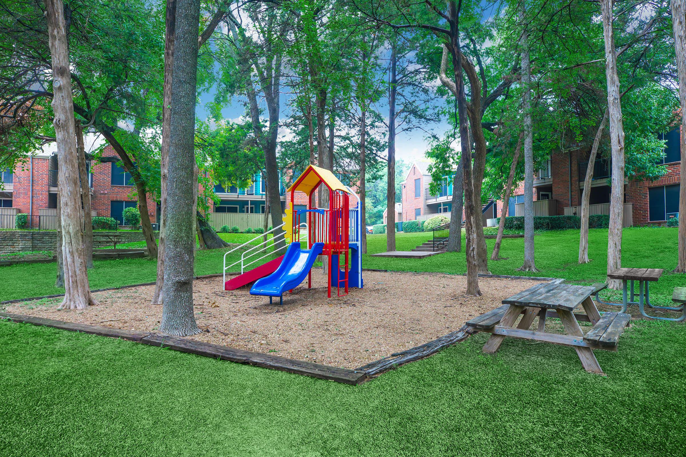A colorful children's playground with a red and yellow play structure featuring slides, surrounded by lush green grass and trees. A picnic table and benches are visible nearby, creating a playful and inviting outdoor space. The area is well-maintained, ideal for family activities and recreation.