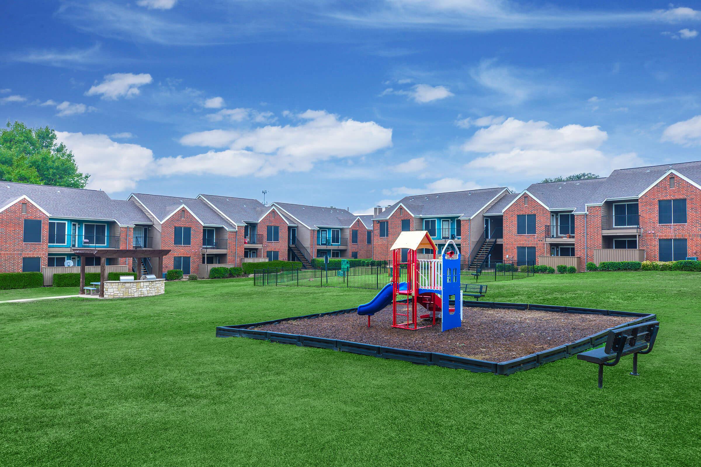 A grassy area featuring a playground with colorful equipment, including slides and climbing structures. Surrounding the playground are benches and two-story brick apartment buildings in the background, with a clear blue sky overhead.