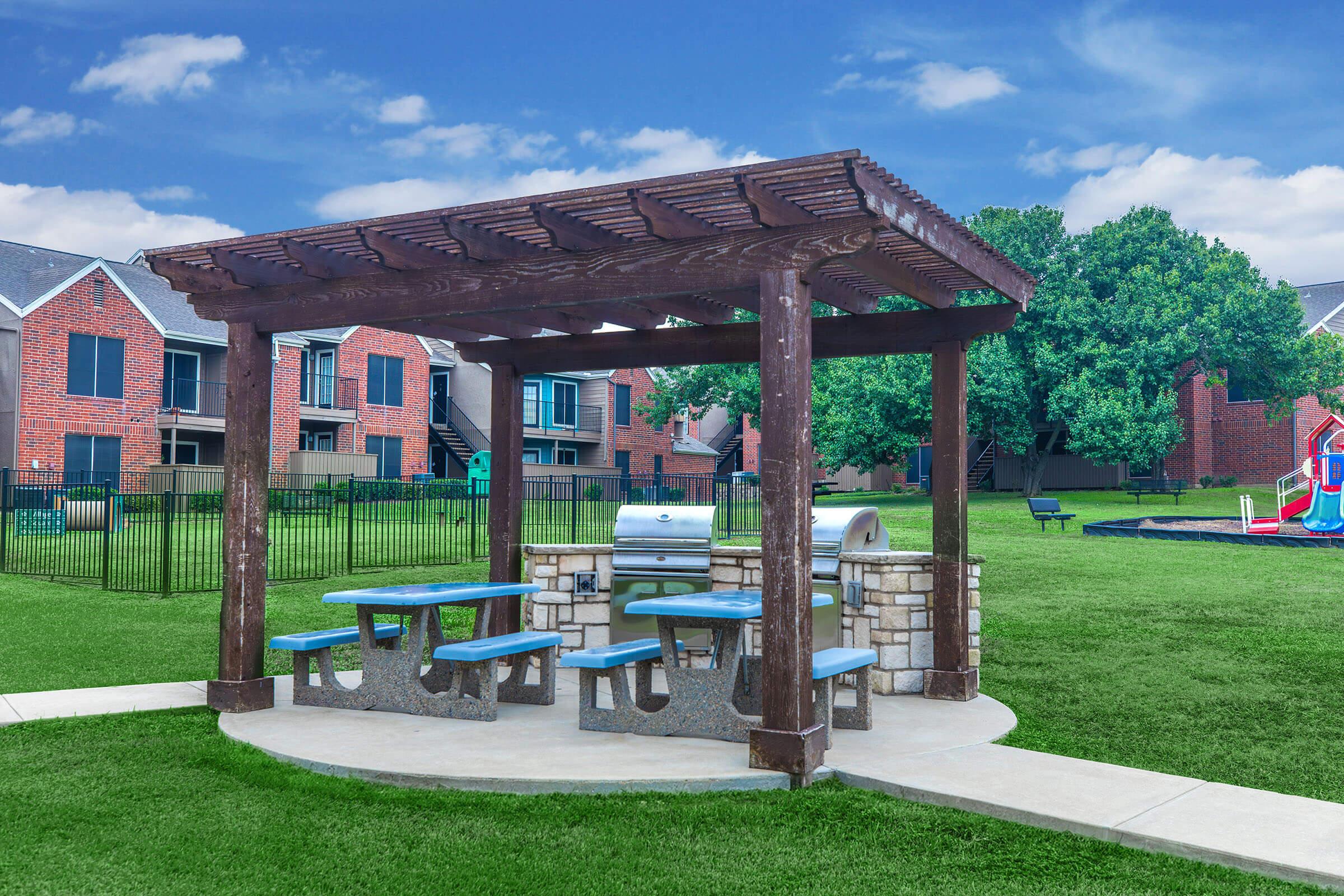 Outdoor picnic area with a wooden pergola and blue picnic tables. A grill is located nearby, surrounded by a grassy space. In the background, there are residential buildings and a playground, creating a family-friendly setting. The sky is clear with clouds, adding to the pleasant atmosphere.