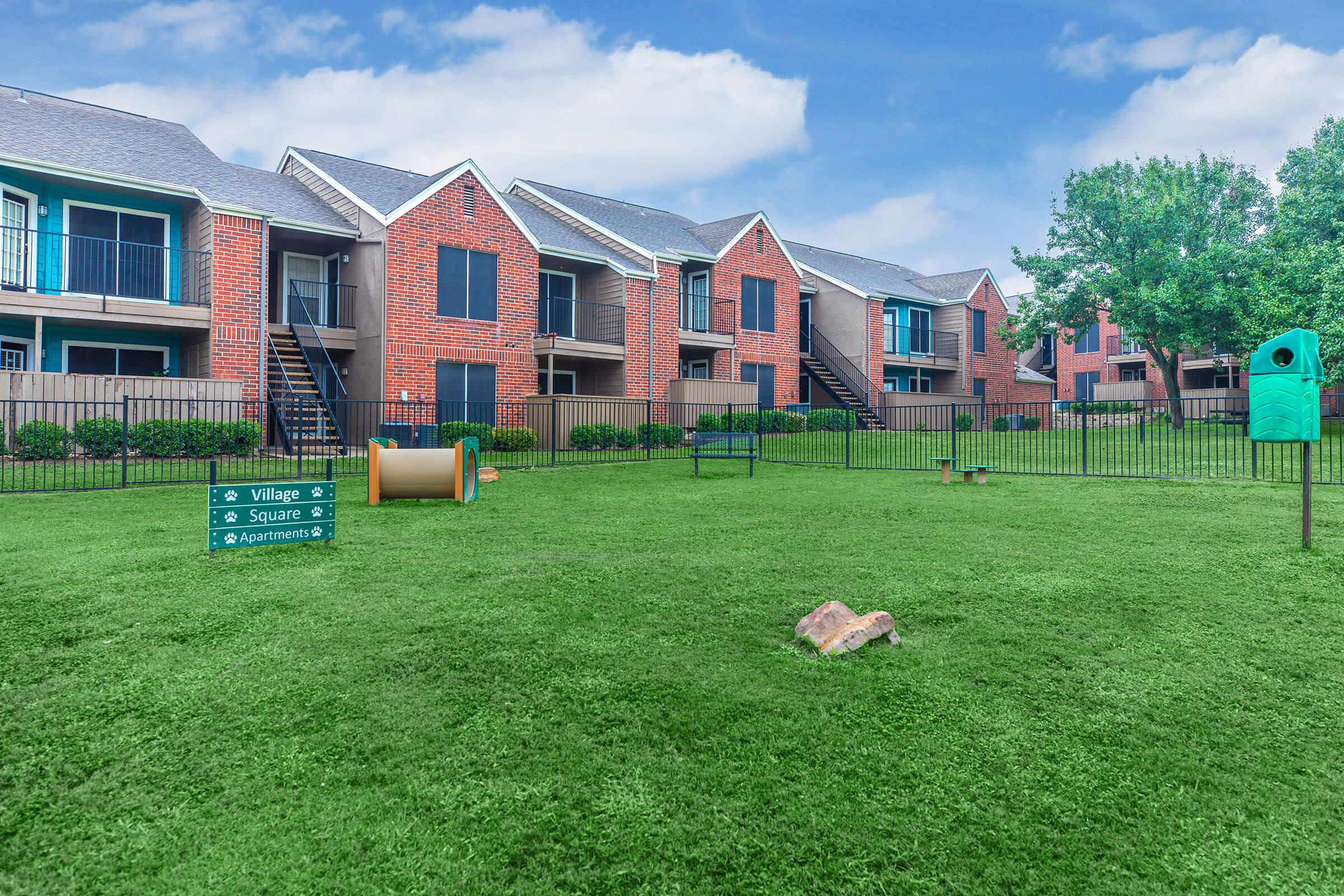 A spacious green lawn in front of a brick apartment complex, featuring multiple buildings with balconies. There are signs for "Village Square Apartments," and a pet area with a tunnel and waste station. The sky is blue with some clouds, and lush shrubbery borders the property.
