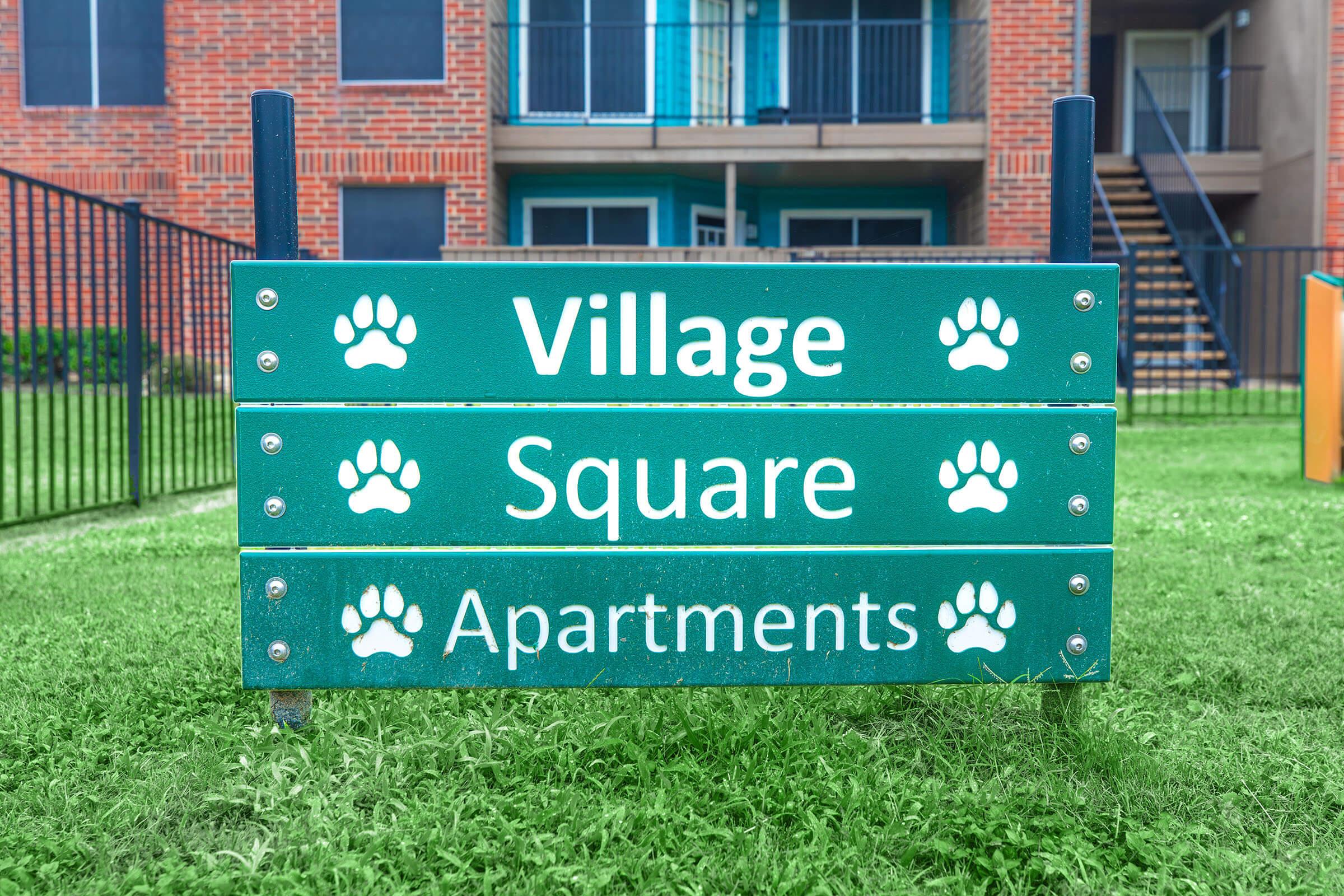 A green sign with white text displaying "Village," "Square," and "Apartments," accompanied by paw print icons on each line, located in a grassy area in front of a brick apartment building with a staircase and railing in the background.