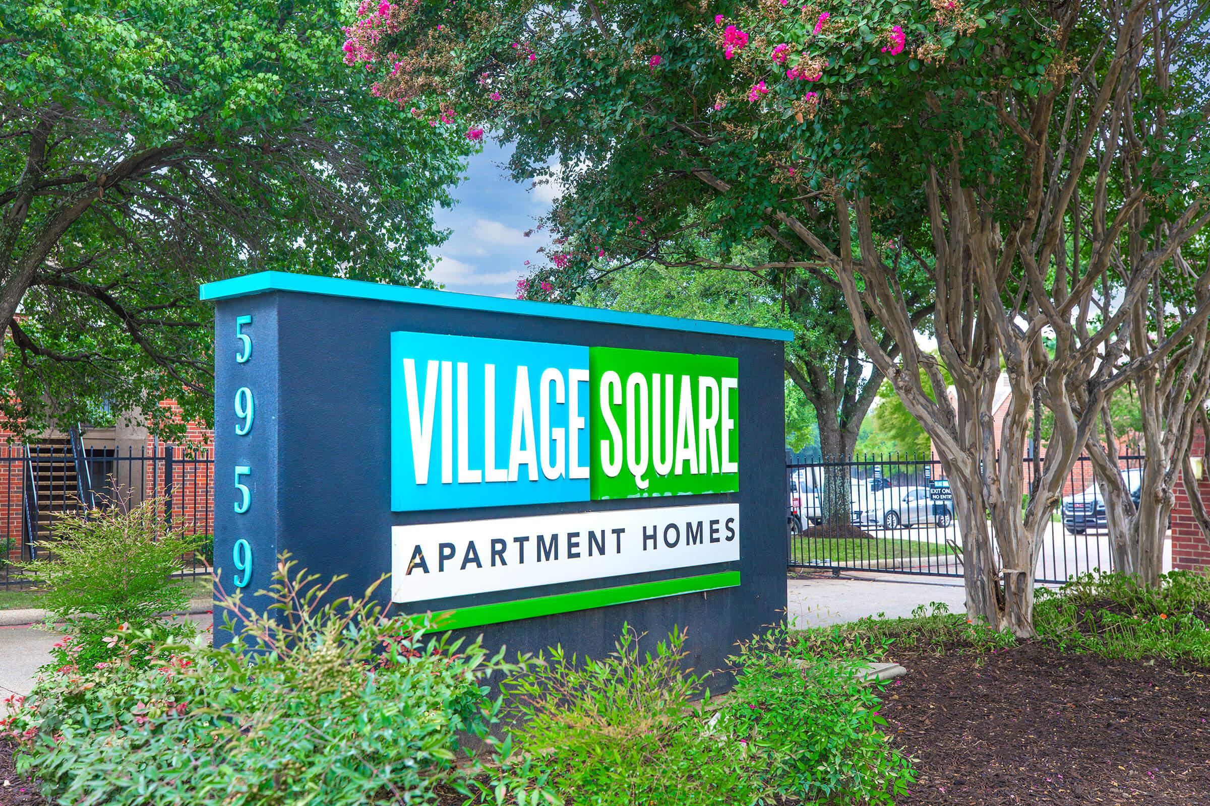 Sign for Village Square Apartment Homes, featuring bright blue and green colors. The sign is surrounded by well-maintained landscaping, including bushes and trees, and is located near a residential area with cars visible in the background. The address, 5959, is displayed prominently on the sign.