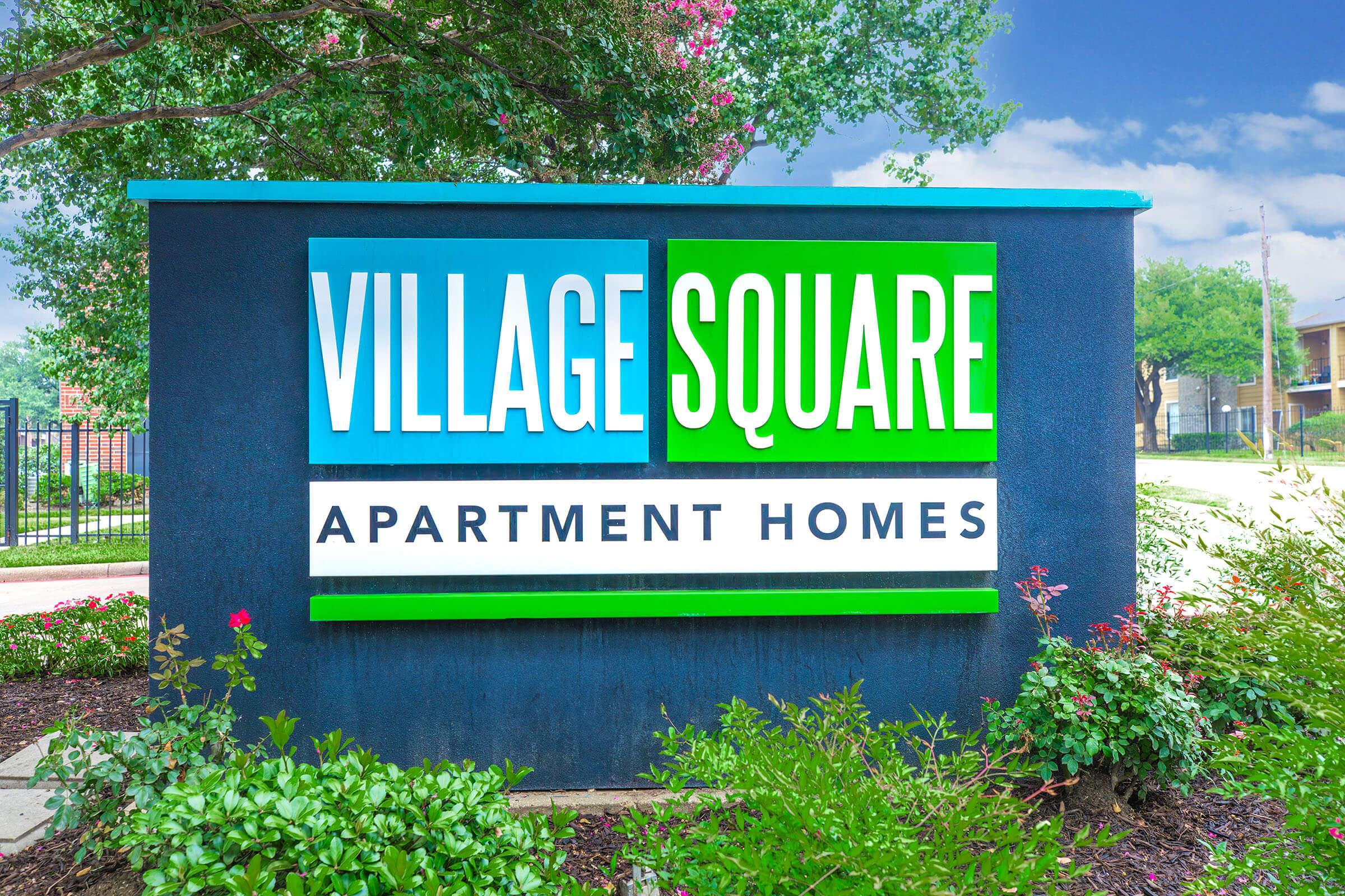 Sign for Village Square Apartment Homes, featuring a blue and green color scheme. The words "VILLAGE" and "SQUARE" are in bold, large letters, while "APARTMENT HOMES" is in smaller text below. The background includes greenery and a clear blue sky.