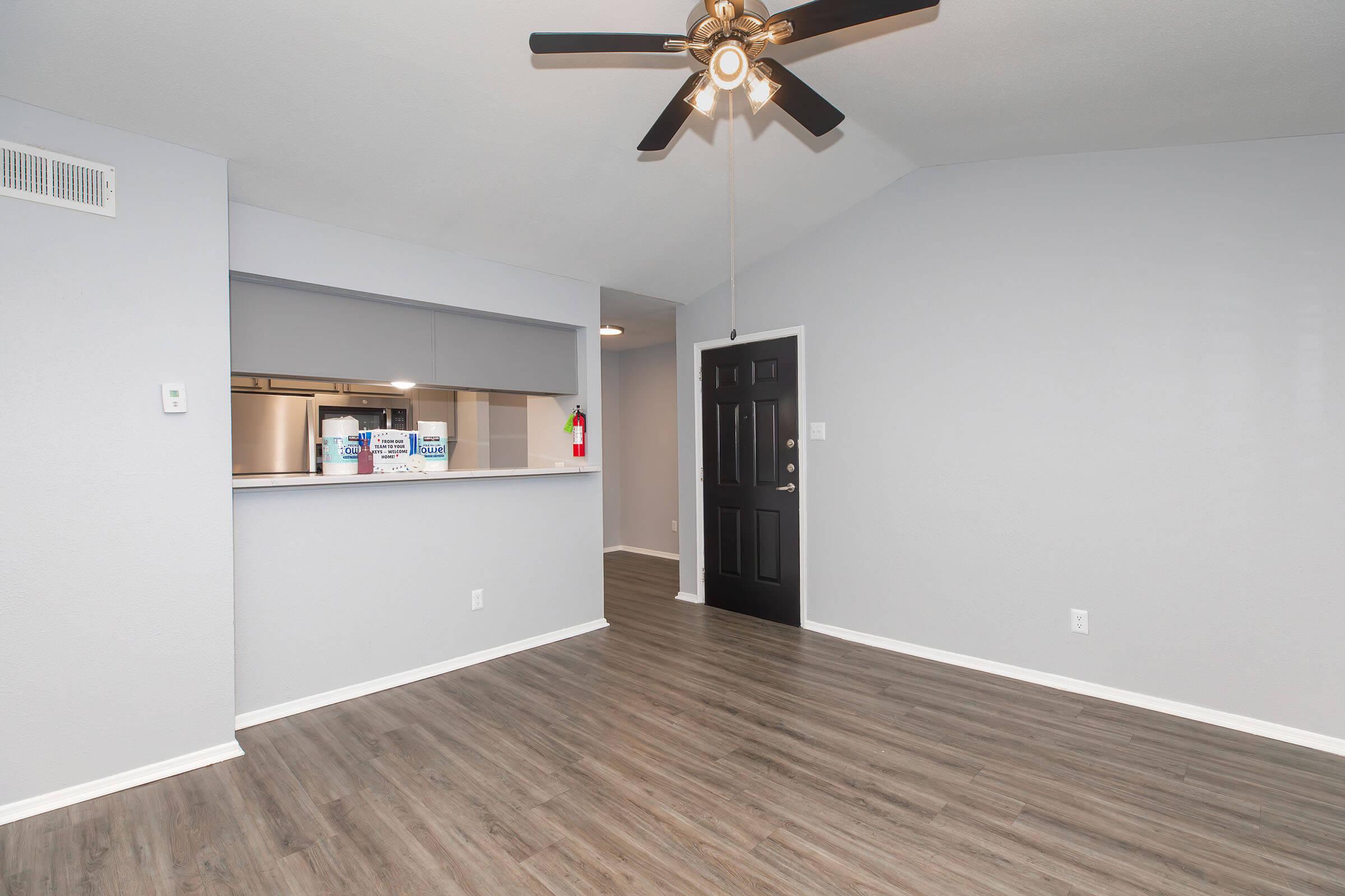 A spacious living area with a ceiling fan, featuring gray walls and hardwood-style flooring. There is an entry door on the right and an open kitchen space in the background with a bar counter. The overall ambiance is modern and well-lit.
