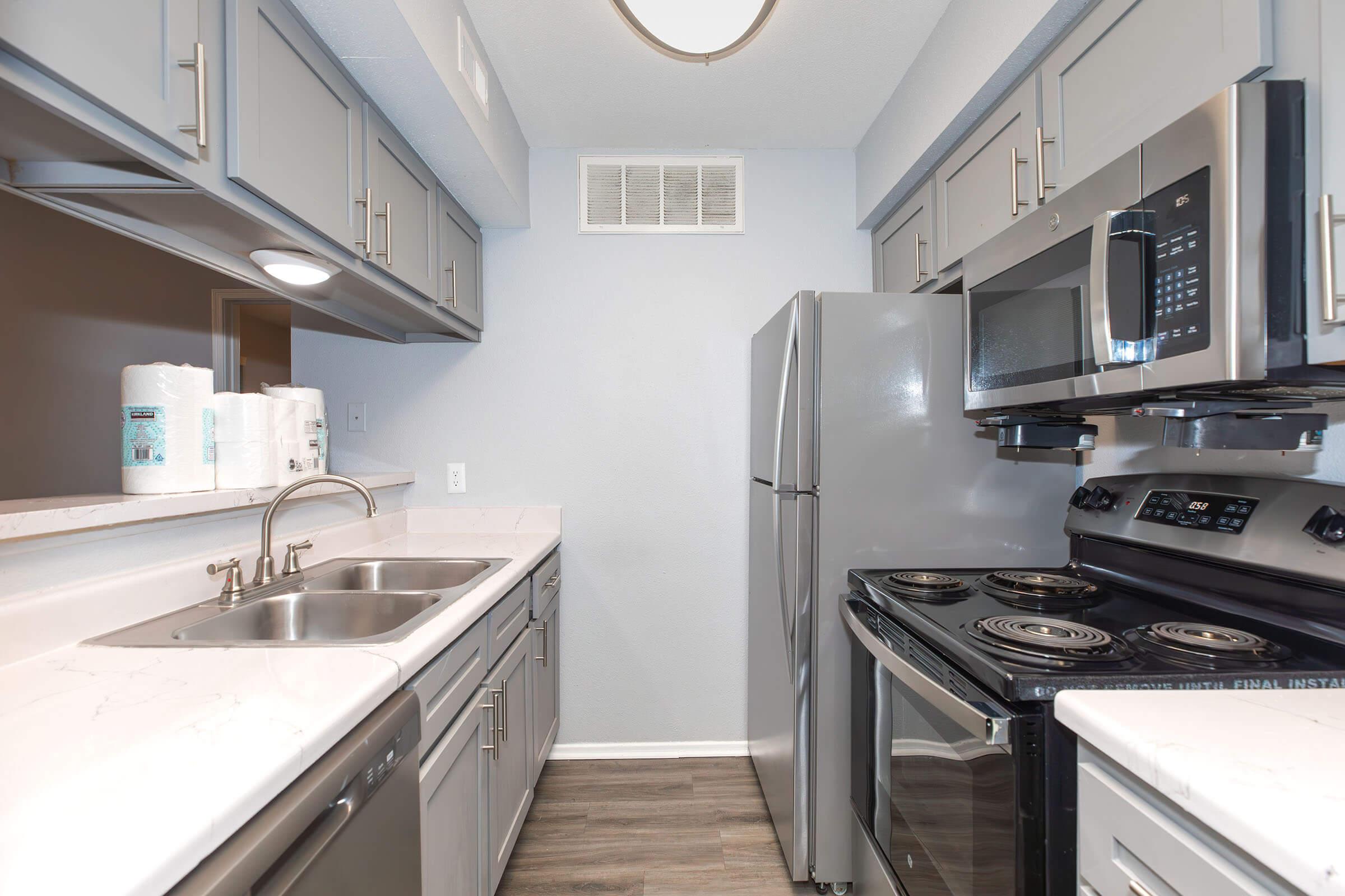 A modern kitchen featuring gray cabinetry, a double sink, stainless steel appliances including a refrigerator, microwave, and stove. The countertops are light-colored, and paper towels are neatly arranged on the counter. The flooring is a wood-like laminate, creating a contemporary look.