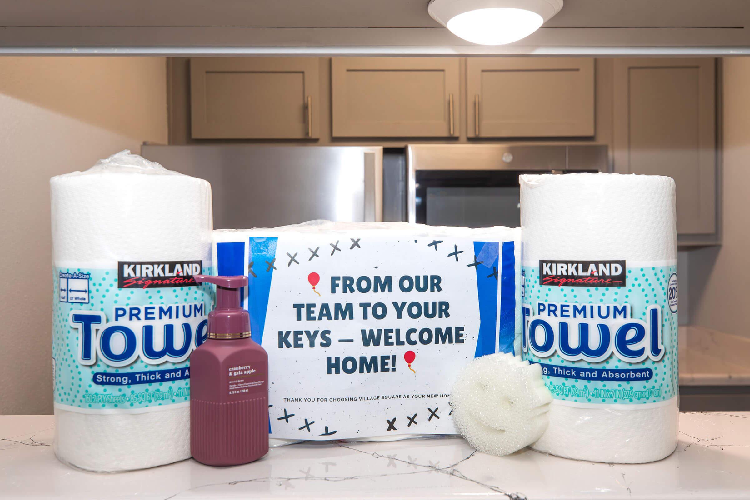 Two rolls of Kirkland brand premium towels are positioned on a kitchen countertop alongside a bottle of lotion and a loofah. In the background, a welcome sign reads, "FROM OUR TEAM TO YOUR KEYS - WELCOME HOME!" indicating a warm welcome for new residents.