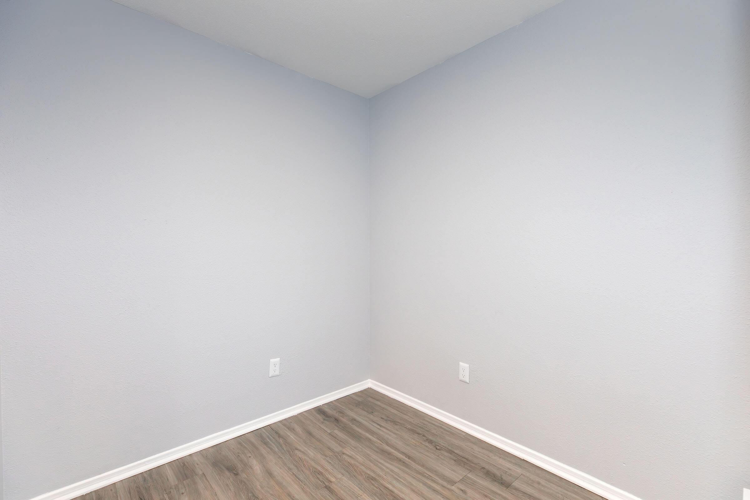 A vacant corner of a room featuring light blue walls and a wooden floor. There are no furnishings or decorations, creating an open and minimalistic space. The walls are smooth, and the corner meets at a 90-degree angle.