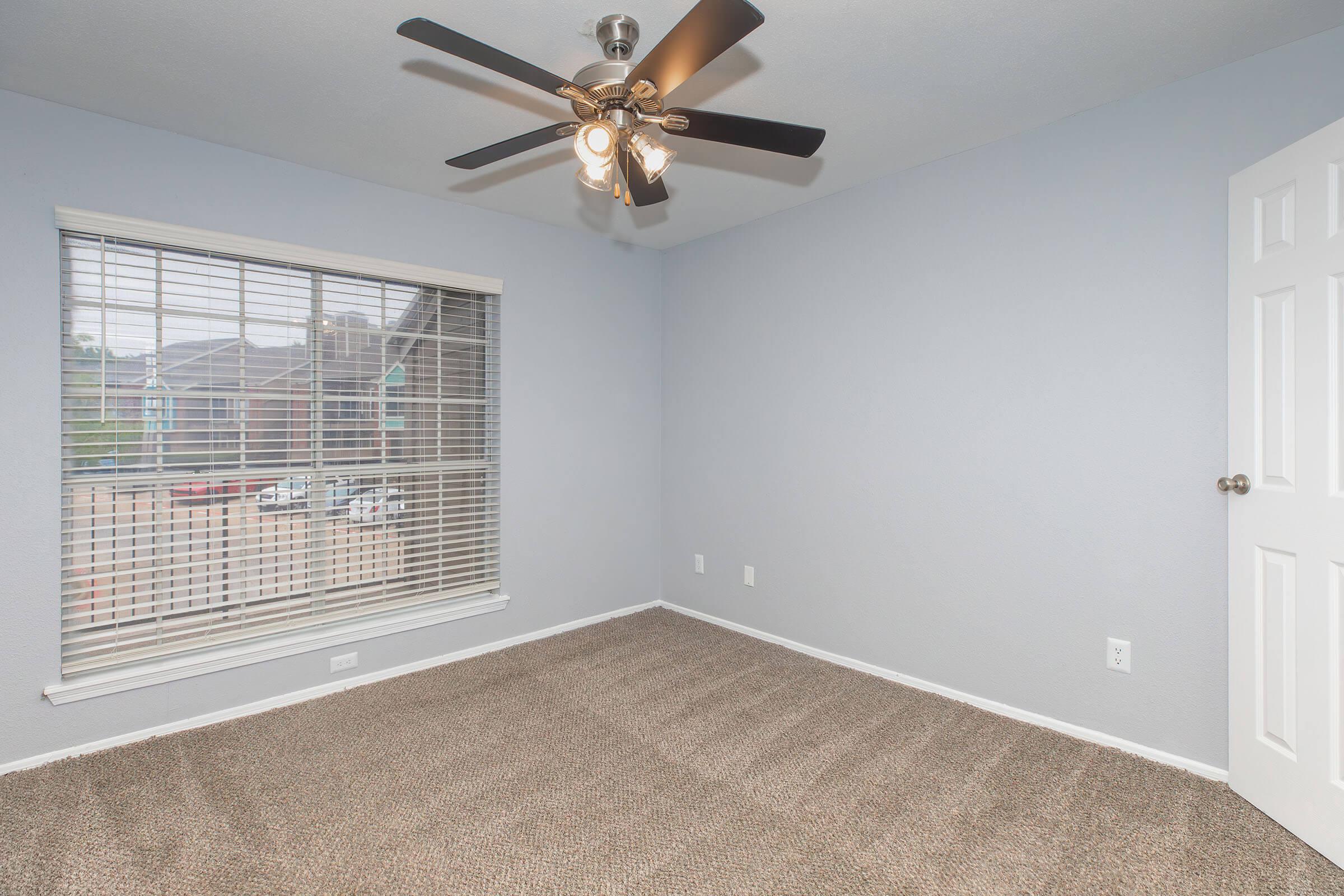 Empty room with light blue walls and a ceiling fan. It has a window with blinds, letting in natural light, and carpeted flooring. The room is unfurnished and appears spacious, creating a clean and simple aesthetic. A door is visible on the right side, leading to another area.