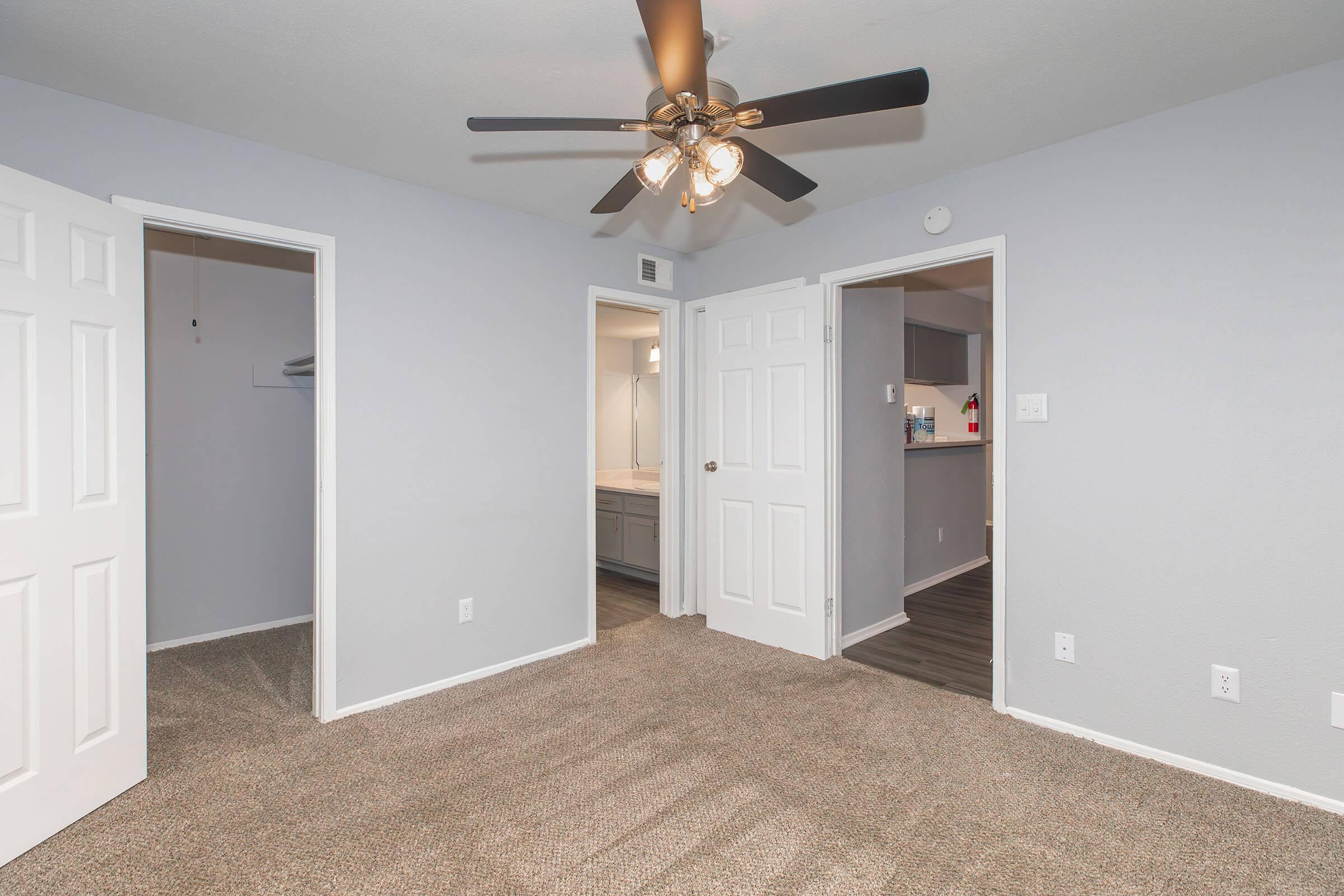 A well-lit room featuring carpeted flooring, a ceiling fan with light, and two white doors leading to different areas. One door is open to a small closet, while the other opens to a bathroom area. The walls are painted a light gray, creating a spacious and inviting atmosphere.