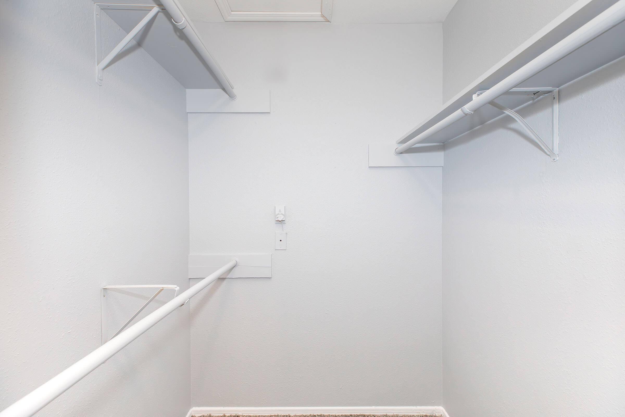 Empty closet with white walls, featuring two white shelves and hanging rods on either side. The floor is bare, and there is a ceiling vent visible. Overall, the closet has a clean and minimalist appearance.