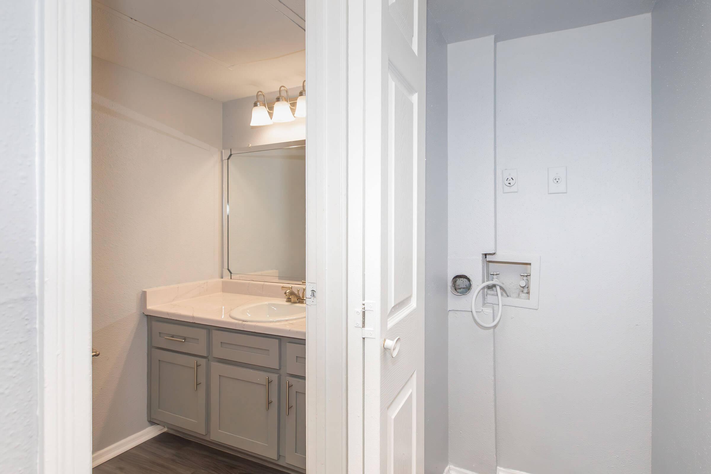 A small bathroom featuring a light gray vanity with a sink and mirror, illuminated by three overhead lights. The walls are painted in a light shade, and there is a door leading to another room. The flooring is a dark laminate, creating a contrast with the walls and fixtures.