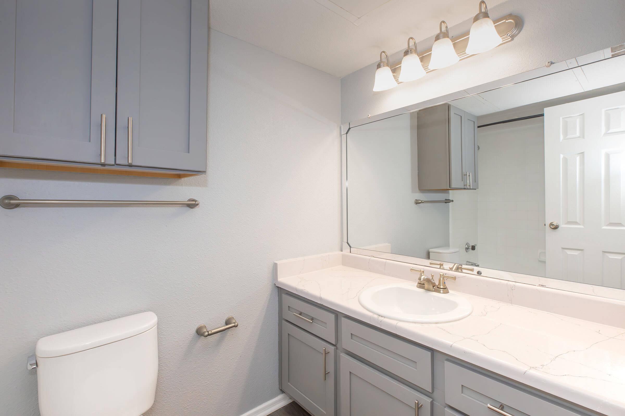 A modern bathroom featuring a light gray vanity with a marble countertop, a round sink, and a gold faucet. Above the vanity, there is a large mirror and a light fixture with three bulbs. The walls are painted in a light color, and a door leads to a shower area. A toilet is positioned to the left of the vanity.