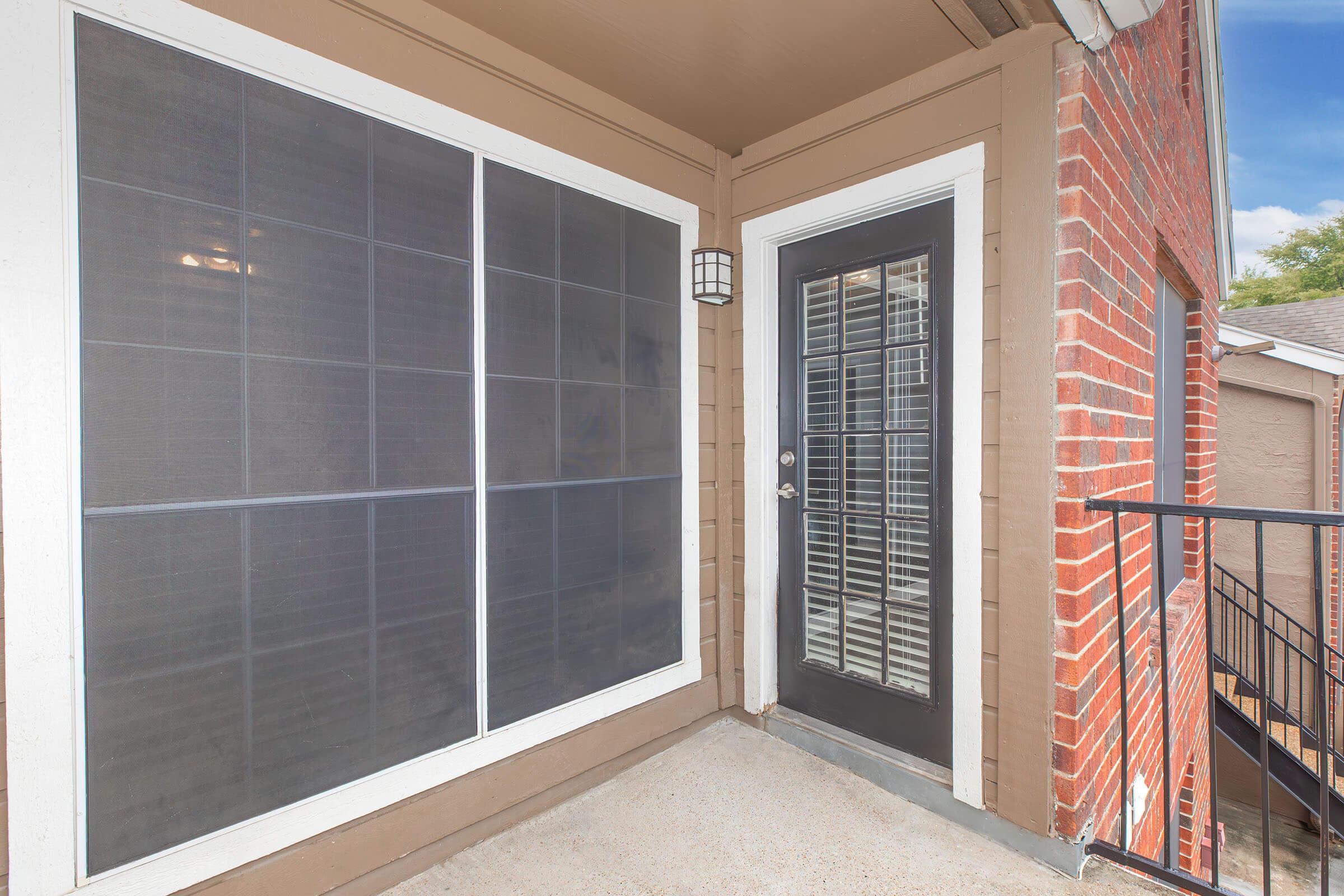 A covered entrance with a black door and windowed panels on the side. The walls are a mix of brown siding and red brick. There's a light fixture next to the door, and a metal railing leads away from the doorway. The porch is empty, with a light blue sky visible in the background.