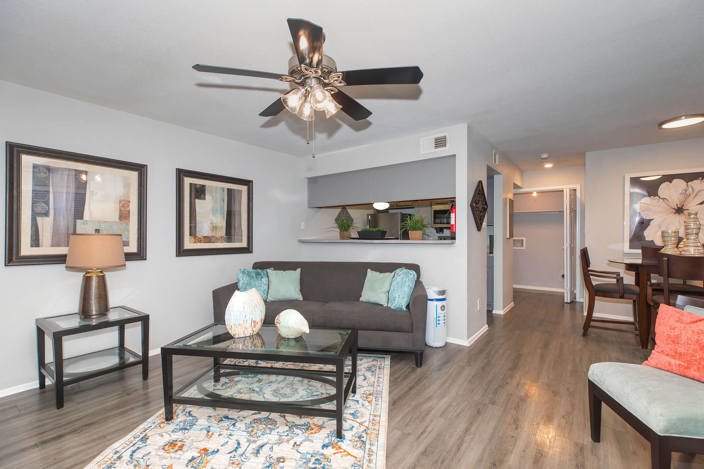 Modern living room featuring a gray sofa with turquoise accent pillows, a glass coffee table on an ornate area rug, and framed artwork on the walls. A ceiling fan with light hangs above, and there is a dining area visible in the background, along with a bright, airy feel throughout the space.