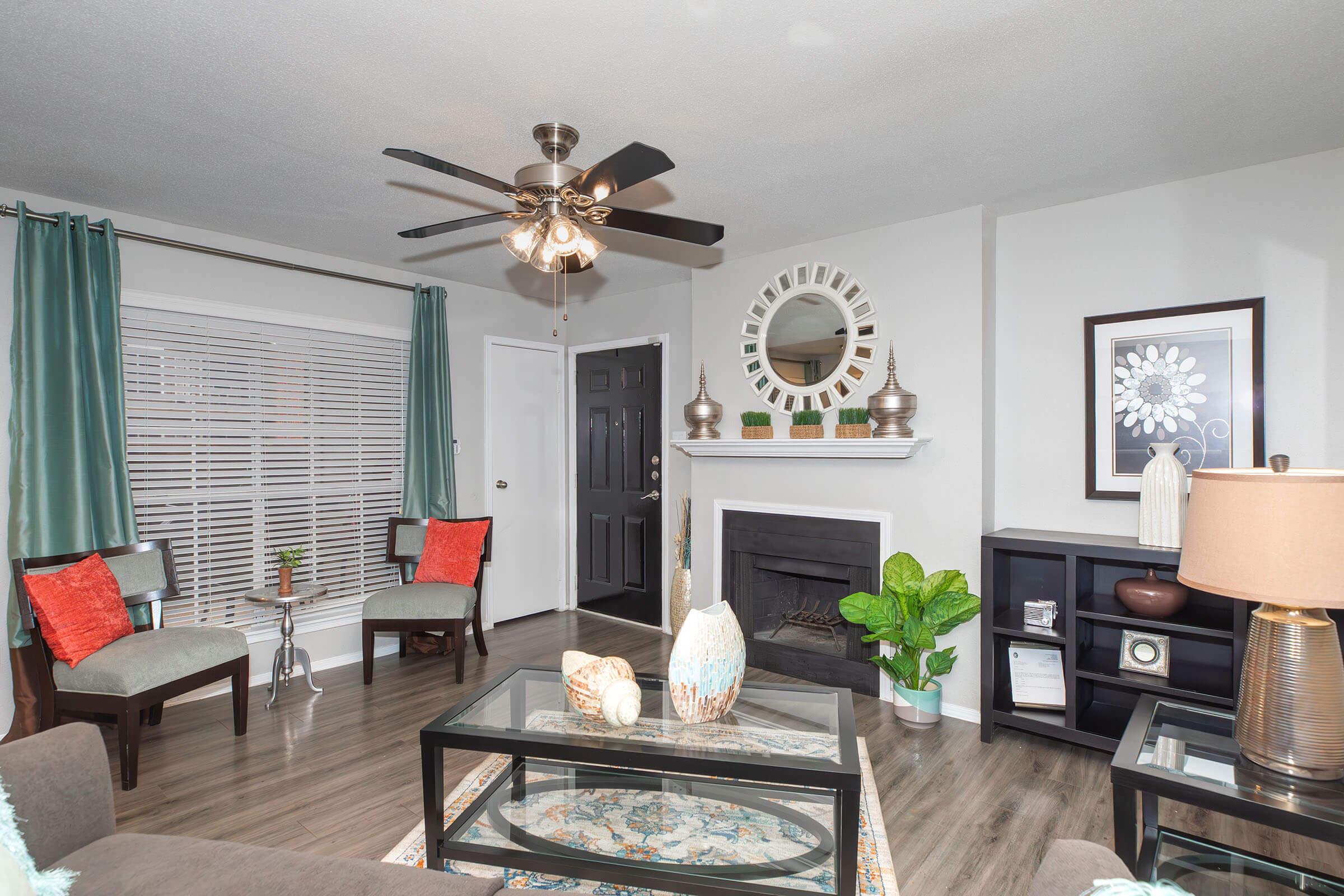 A cozy living room featuring a ceiling fan, two chairs with orange cushions, and a decorative coffee table. There's a fireplace with a circular mirror above it, and a plant in the corner. Adjacent to the fireplace are shelves displaying decorative items, and large windows covered with green curtains.