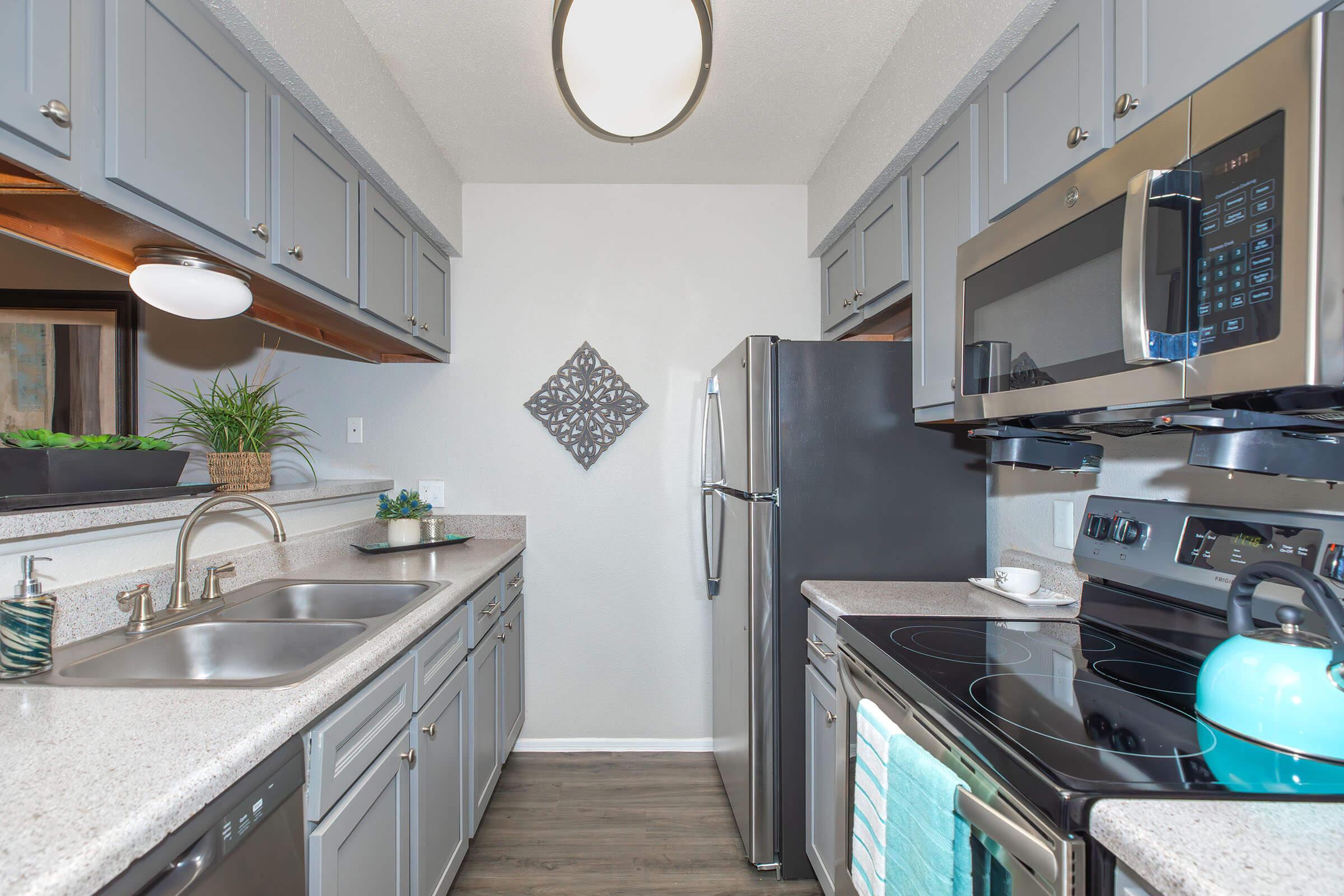 A modern kitchen featuring gray cabinets, stainless steel appliances, and a double sink. The countertop is light-colored with a decorative plant. On the wall, there's a simple wall art piece. Natural light enters through a window, enhancing the clean and contemporary look of the space.