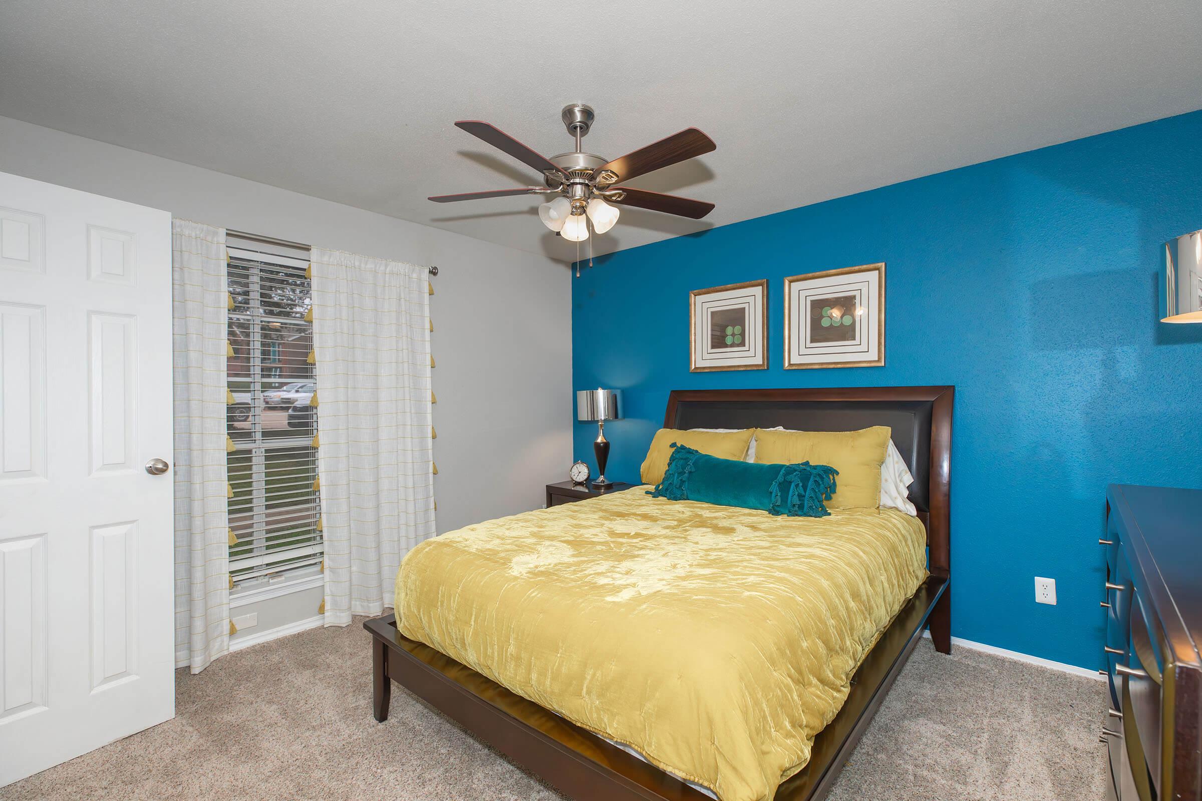 Cozy bedroom featuring a queen-sized bed with a yellow comforter and decorative pillows, a wooden bed frame, nightstands with lamps, and a bright blue accent wall. A ceiling fan hung from the ceiling complements the room's modern look, and a window with sheer curtains allows natural light.