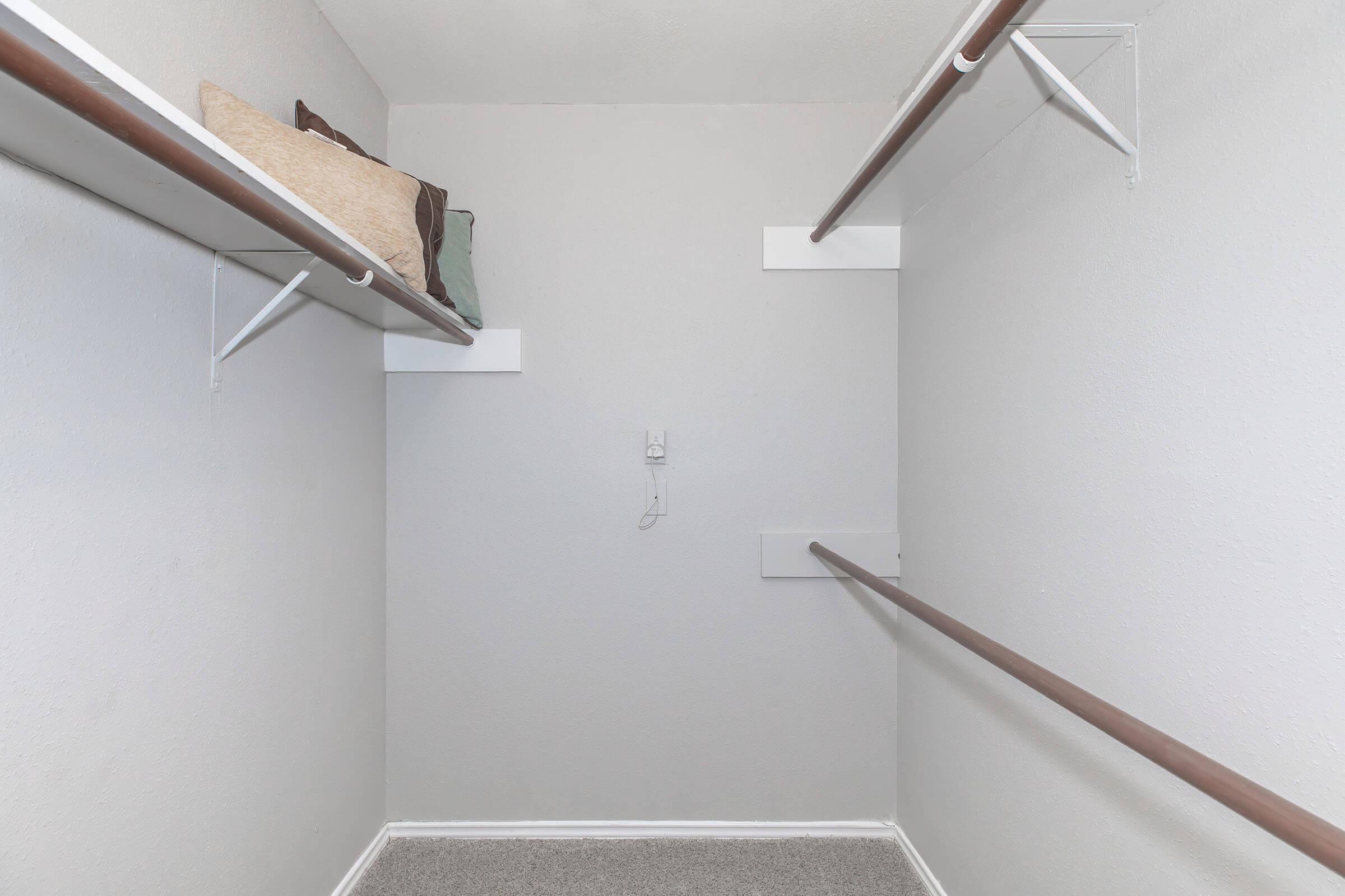 A small, empty closet with light gray walls and carpeted flooring. It features two horizontal clothing rods mounted on the walls and a shelf with a few decorative pillows on the left side. The space appears clean and organized, lacking any visible clothing or items.