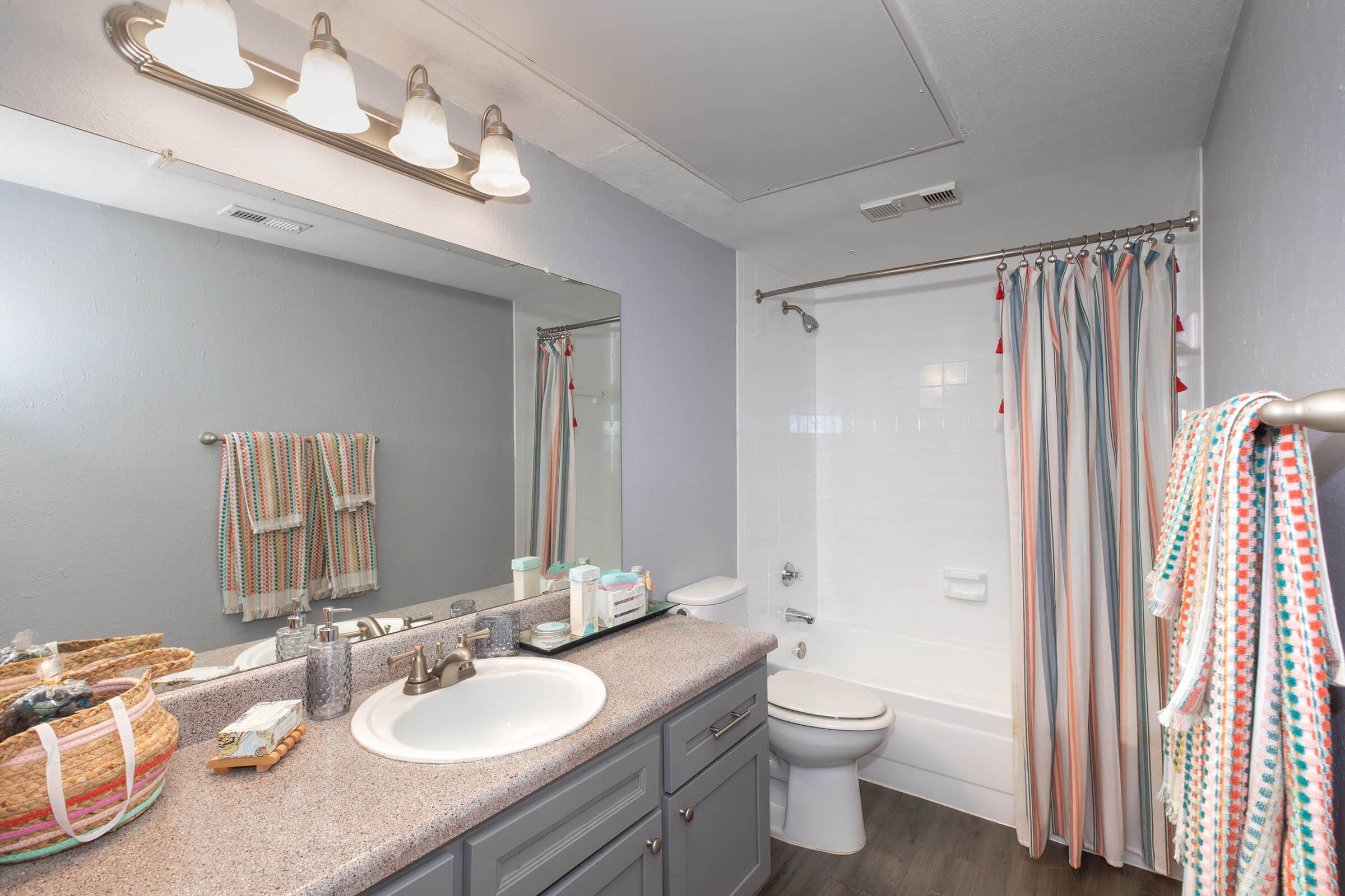 A well-lit bathroom featuring a large mirror above a double sink vanity, with a light gray color scheme. The bathtub is enclosed with a colorful striped shower curtain, and there are neatly organized toiletries and accessories on the counter. Towels hang beside the shower.
