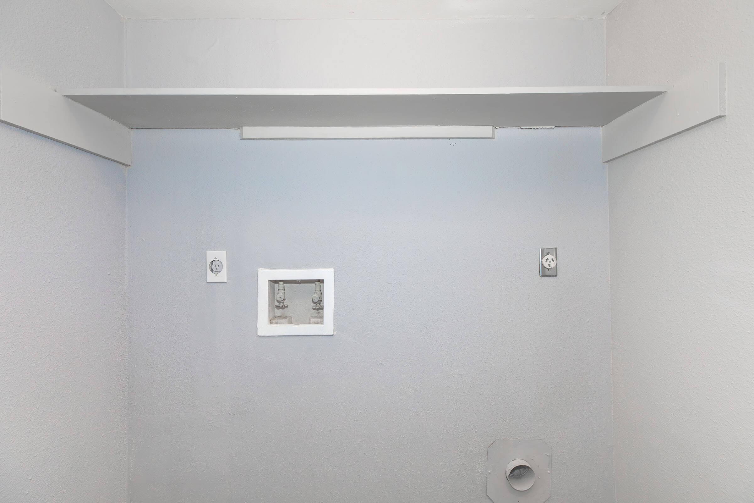 A simple room with gray walls featuring a white shelf at the top. There are two electrical outlets on the left side and a small white panel on the wall. A circular opening is visible at the bottom right, suggesting possible plumbing or wiring access. The overall aesthetic is minimalistic and clean.
