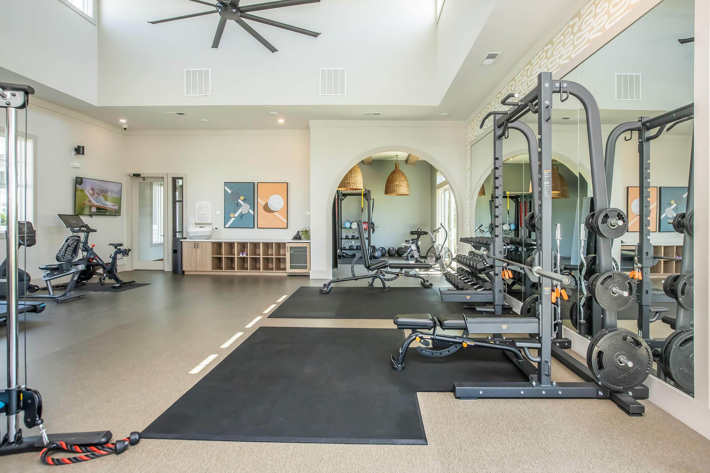 A modern gym interior featuring various exercise equipment, including weights, a bench, a treadmill, and an elliptical machine. The space is well-lit with large windows, sleek flooring, and decorative wall art, creating an inviting atmosphere for fitness activities.