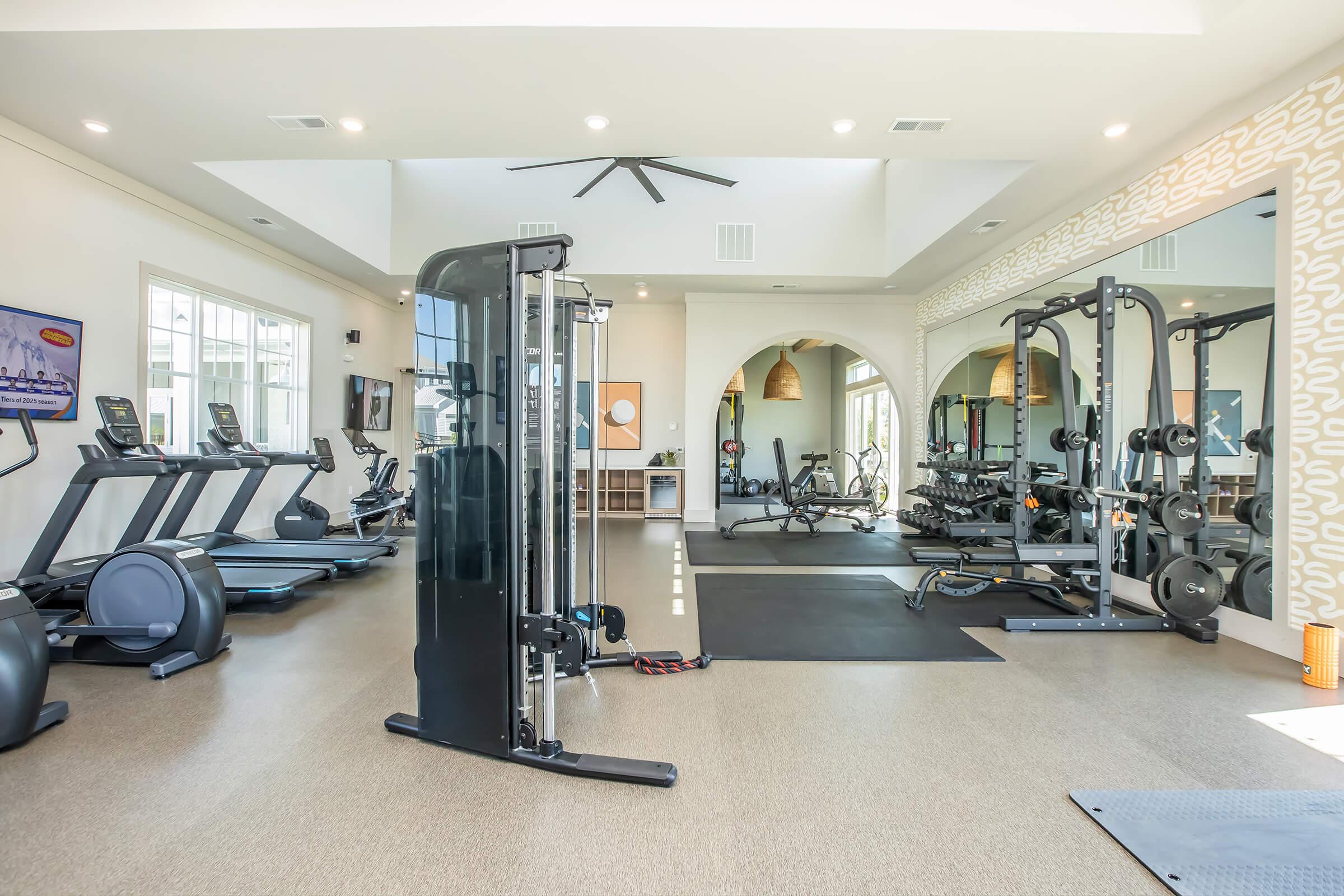 A bright and modern gym with various exercise equipment, including treadmills, stationary bikes, weight machines, and free weights. Large windows let in natural light, and there's an organized layout with mats on the floor, creating an inviting workout space.