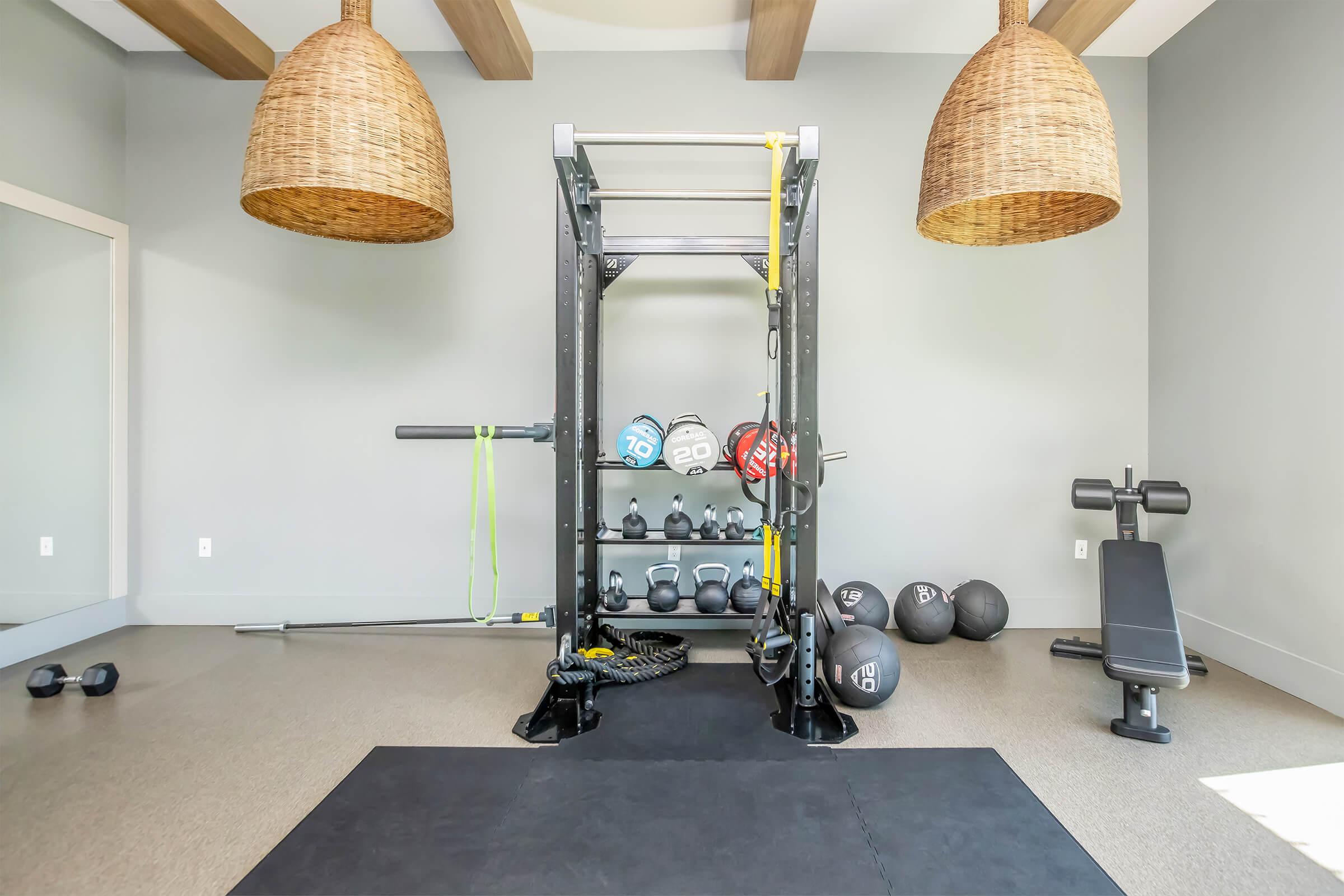 A modern home gym featuring a power rack with weight plates, dumbbells on a rack, a resistance band, and a flat weight bench. The room has light gray walls, wooden beams on the ceiling, and two large woven pendant lights hanging above. A black exercise mat is positioned on the floor.