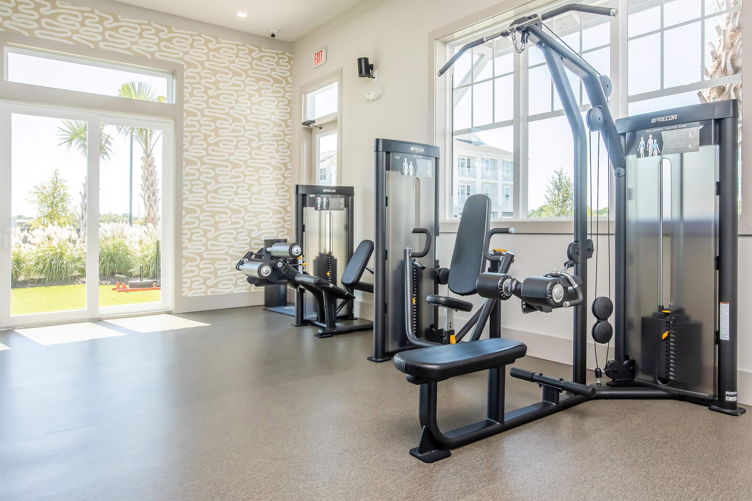 A modern gym interior featuring several exercise machines, including a weight machine and benches. Large windows allow natural light, and there are views of greenery outside. The walls have a stylish patterned design, creating an inviting workout space.