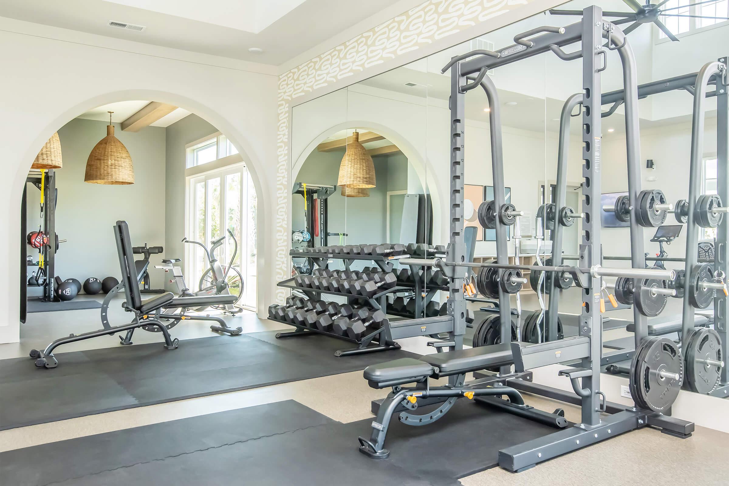 A modern gym interior featuring a variety of fitness equipment. The space includes weight benches, free weights on racks, and exercise machines, all set against a backdrop of large windows. Natural light floods the room, highlighting the clean, airy design with warm pendant lights.