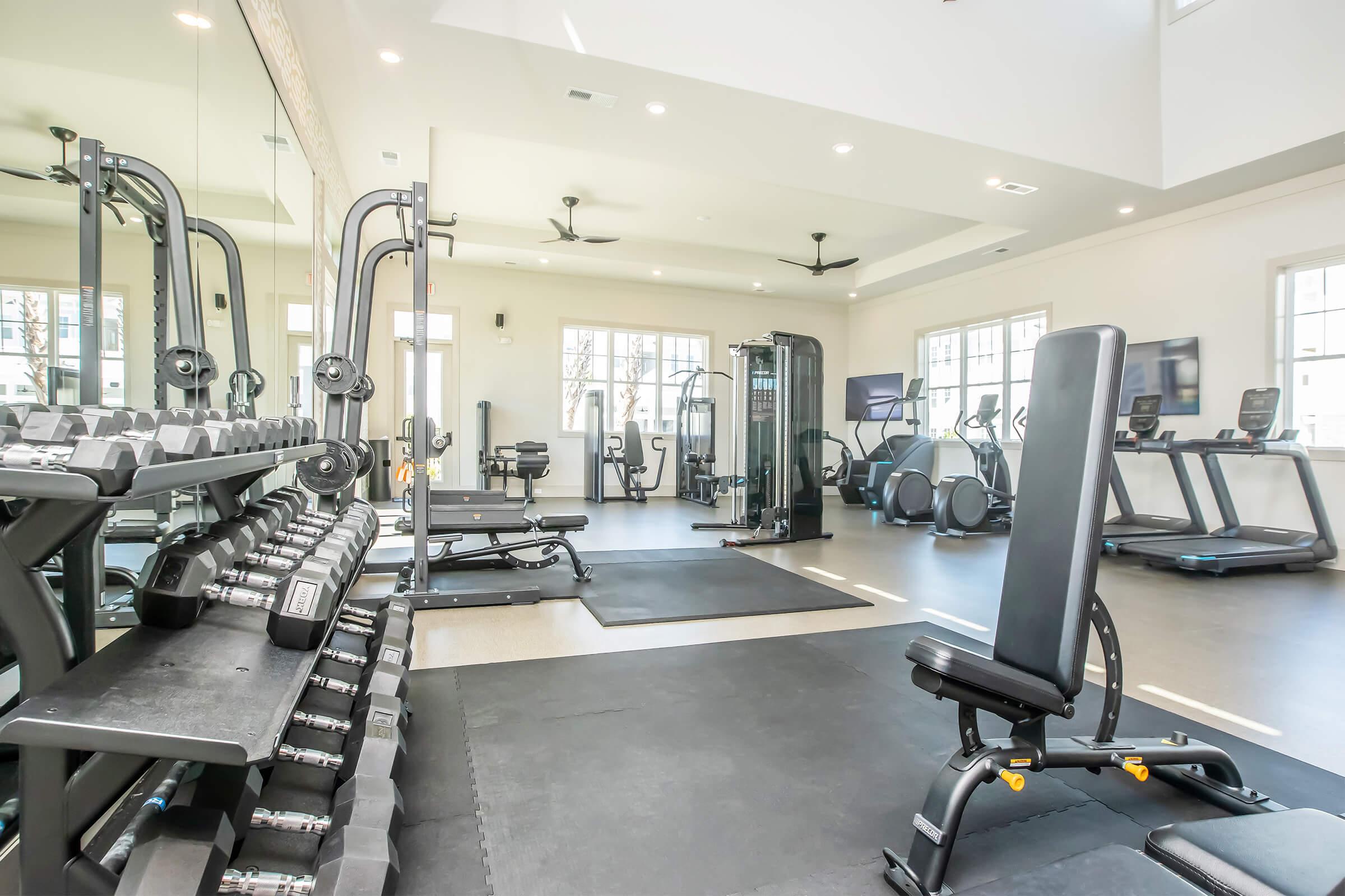 A modern gym interior featuring a range of workout equipment including dumbbells, a weight bench, exercise machines, and treadmills. The space is well-lit with large windows and has a clean, spacious layout designed for fitness activities.