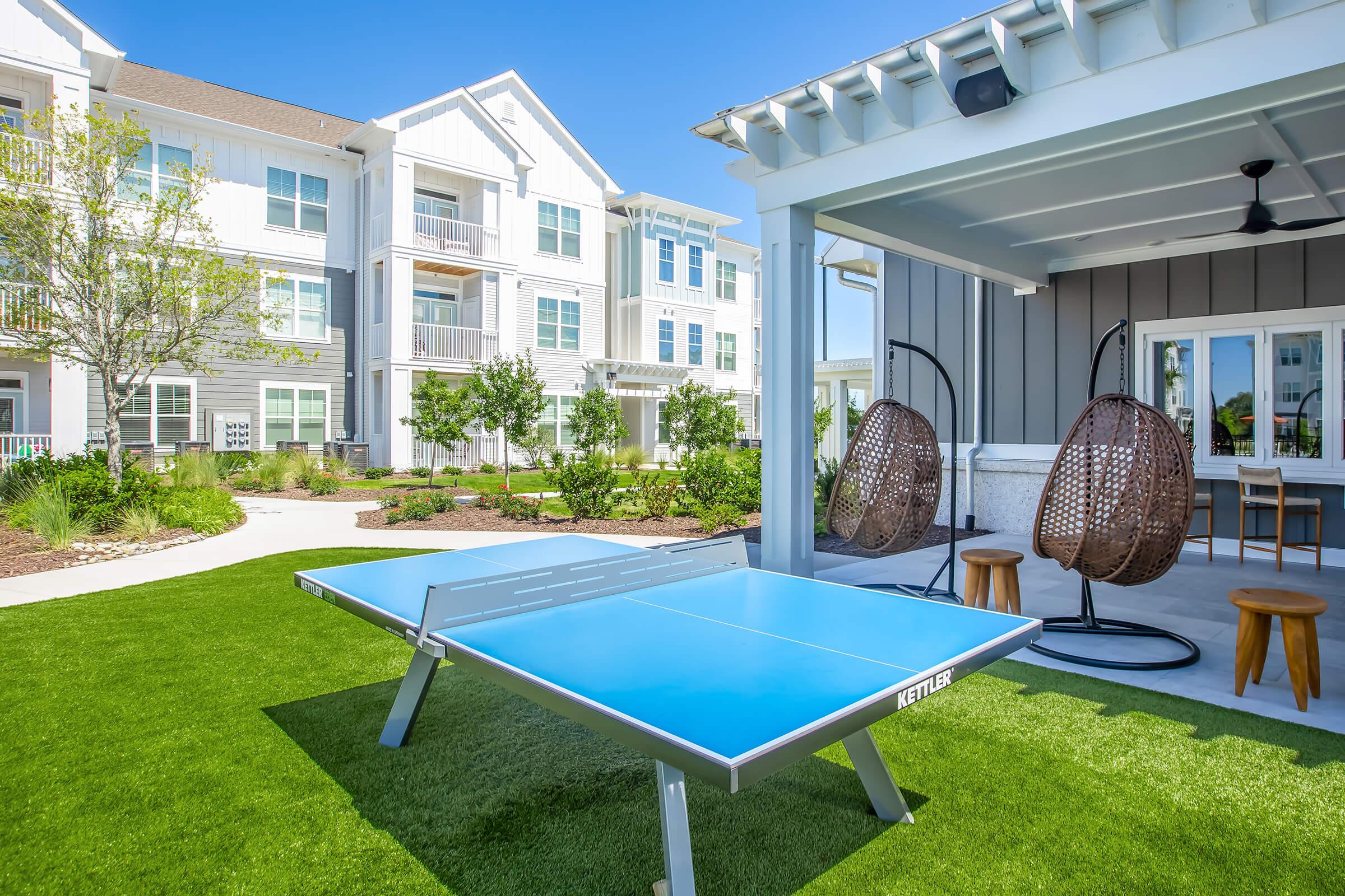 A bright outdoor recreational area featuring a blue ping pong table, two hanging chairs, and a shaded seating area. Lush green grass and modern apartment buildings are visible in the background, creating a vibrant, inviting environment for relaxation and play.
