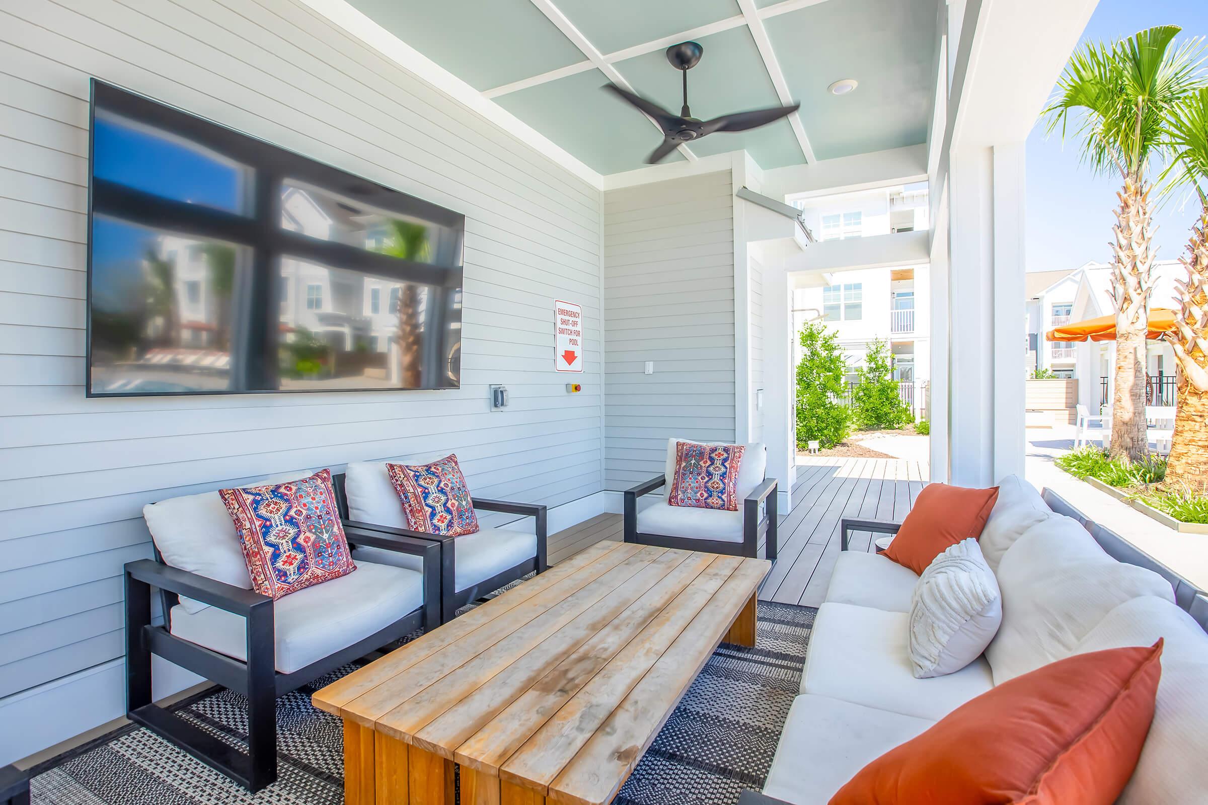 A stylish outdoor lounge area features a comfortable seating arrangement with colorful cushions, a wooden coffee table, and a large wall-mounted television. The space is surrounded by light gray walls, with palm trees visible outside, creating a relaxing atmosphere.