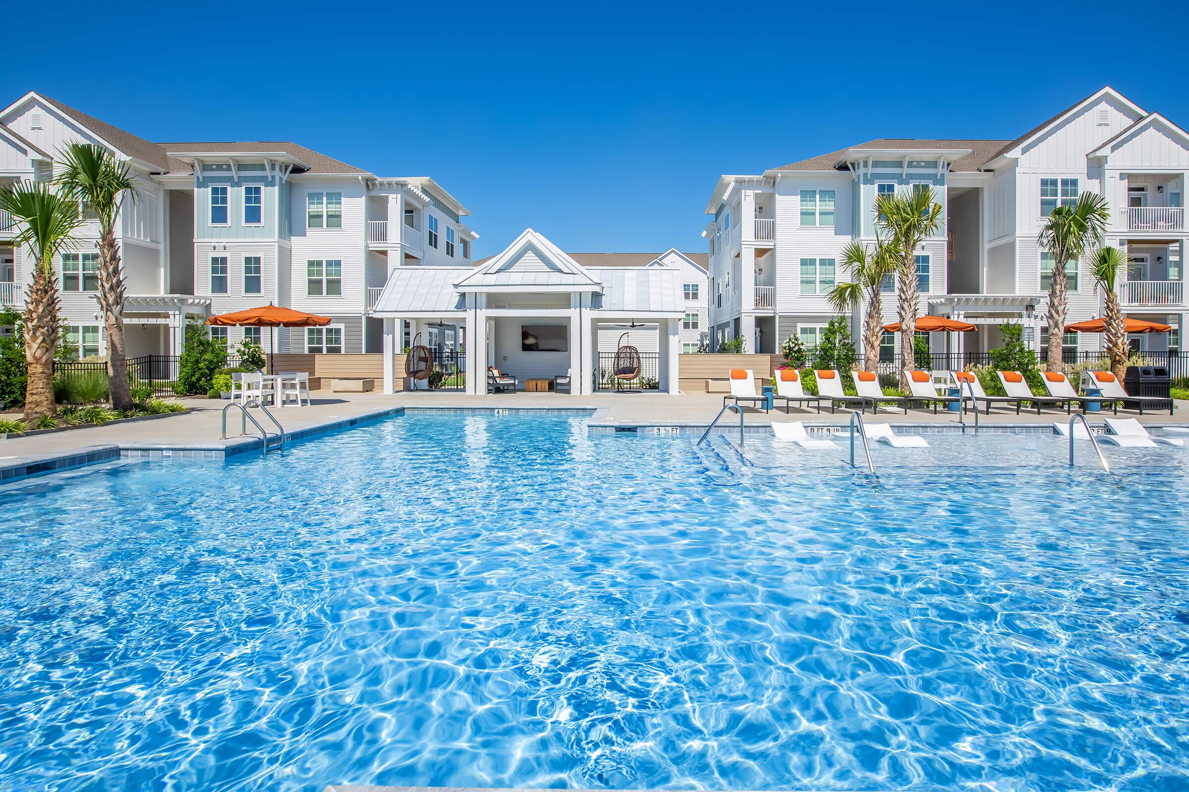 A clear blue swimming pool surrounded by lounge chairs and umbrellas, with palm trees in the background. Modern residential buildings are visible on either side of the pool, under a bright blue sky. The scene conveys a relaxing and inviting atmosphere.