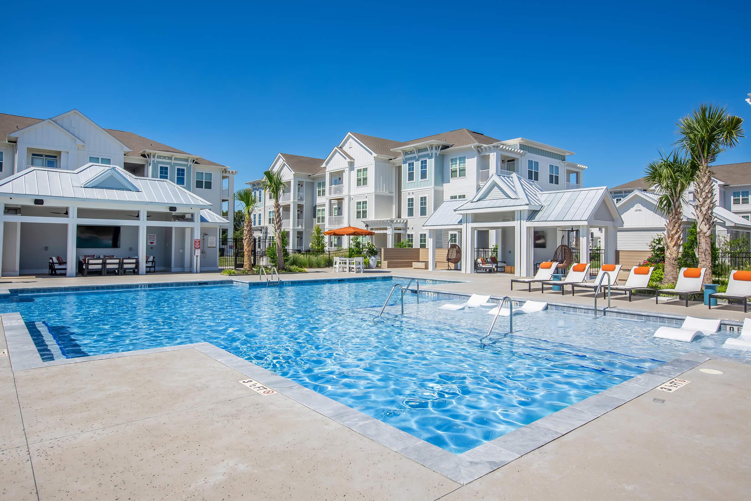 A sparkling blue swimming pool surrounded by lounge chairs and palm trees, with residential buildings in the background. The sky is clear and sunny, creating a relaxing outdoor atmosphere. A shaded area with seating and an umbrella is visible near the poolside.