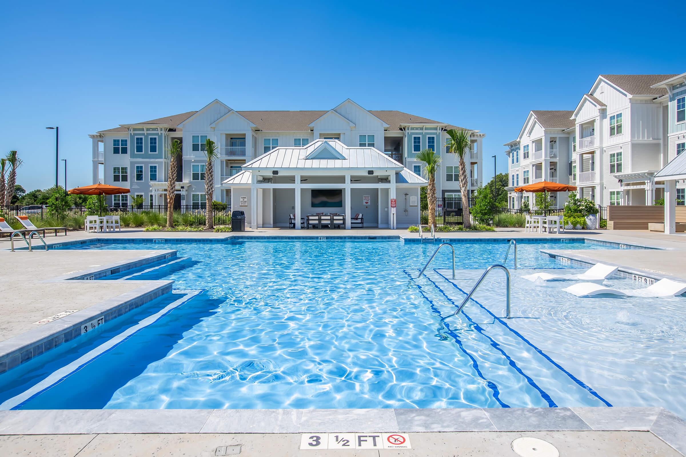 A clear blue swimming pool in a modern residential complex, surrounded by lounge chairs and umbrellas. In the background, a multi-story building with white and light-colored exteriors is visible under a bright blue sky. The pool area is well-maintained and inviting.