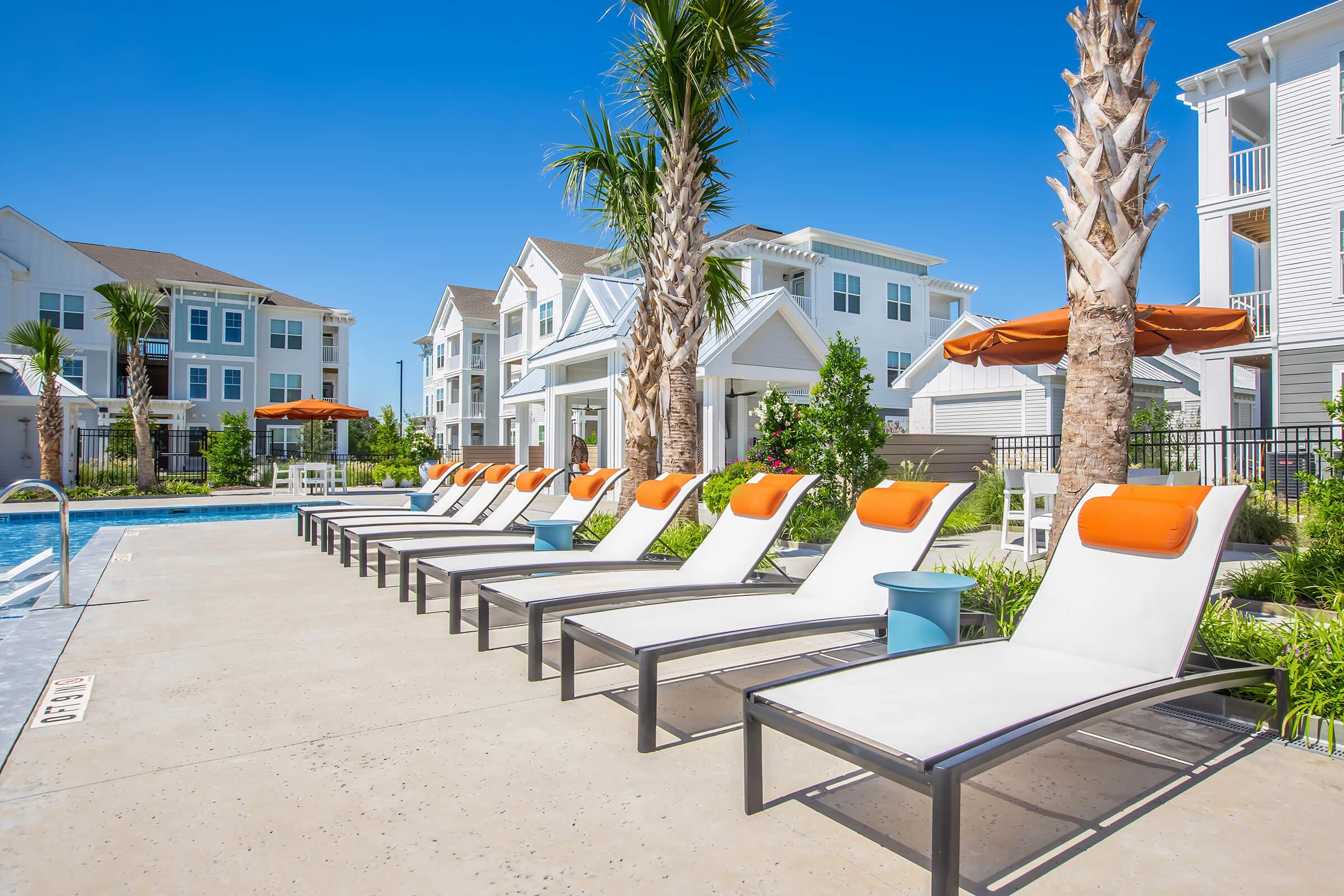A sunny outdoor pool area featuring several lounge chairs with orange cushions, palm trees, and well-maintained landscaping. There are modern buildings in the background and a clear blue sky, creating a vibrant and inviting atmosphere.