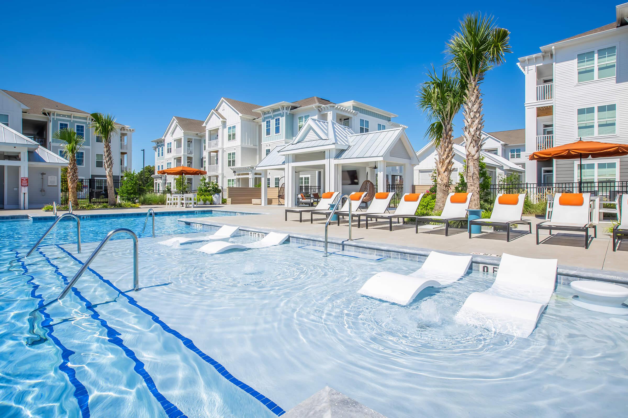A sparkling outdoor swimming pool with lounge chairs featuring orange cushions. Palm trees surround the area, and modern apartment buildings are visible in the background under a clear blue sky. The pool area is well-lit and inviting, perfect for relaxation and leisure activities.