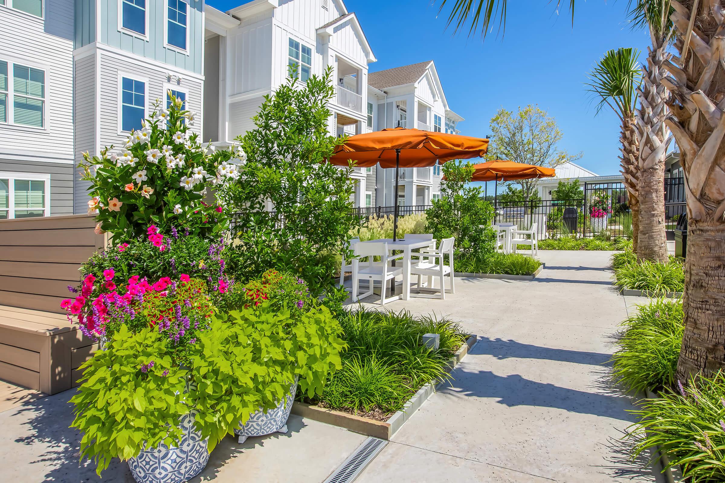 A sunny outdoor area featuring vibrant flower pots with colorful blooms, surrounded by green plants. There are white patio tables and chairs under orange umbrellas, leading to a landscaped pathway and an apartment building in the background. Palm trees add to the tropical ambiance.