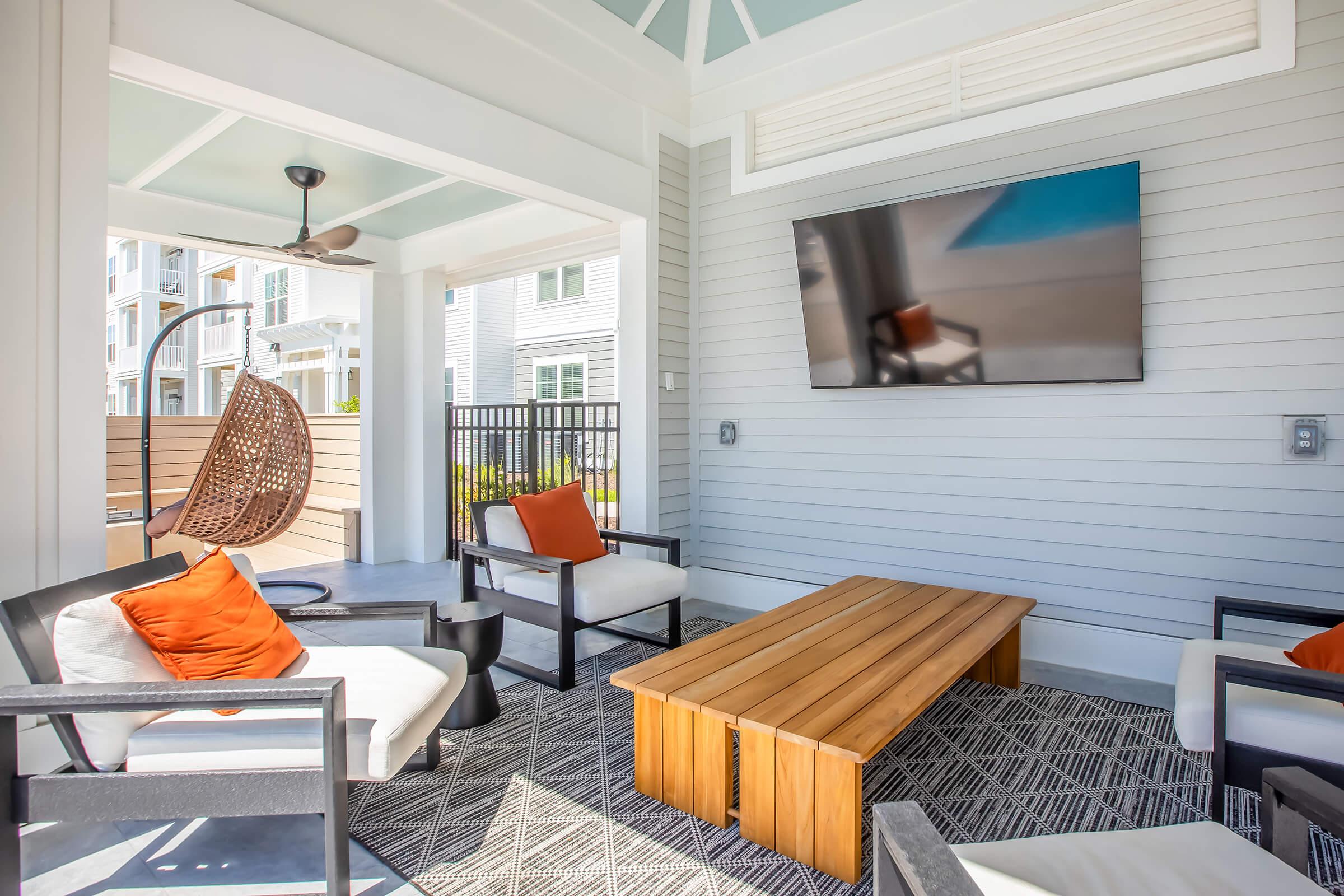 A cozy outdoor lounge area featuring a wooden coffee table, two white chairs with orange cushions, and a hanging chair. A flat-screen TV is mounted on the wall, and the space is surrounded by light-colored wood and modern architecture, with a glimpse of a pool area visible in the background.